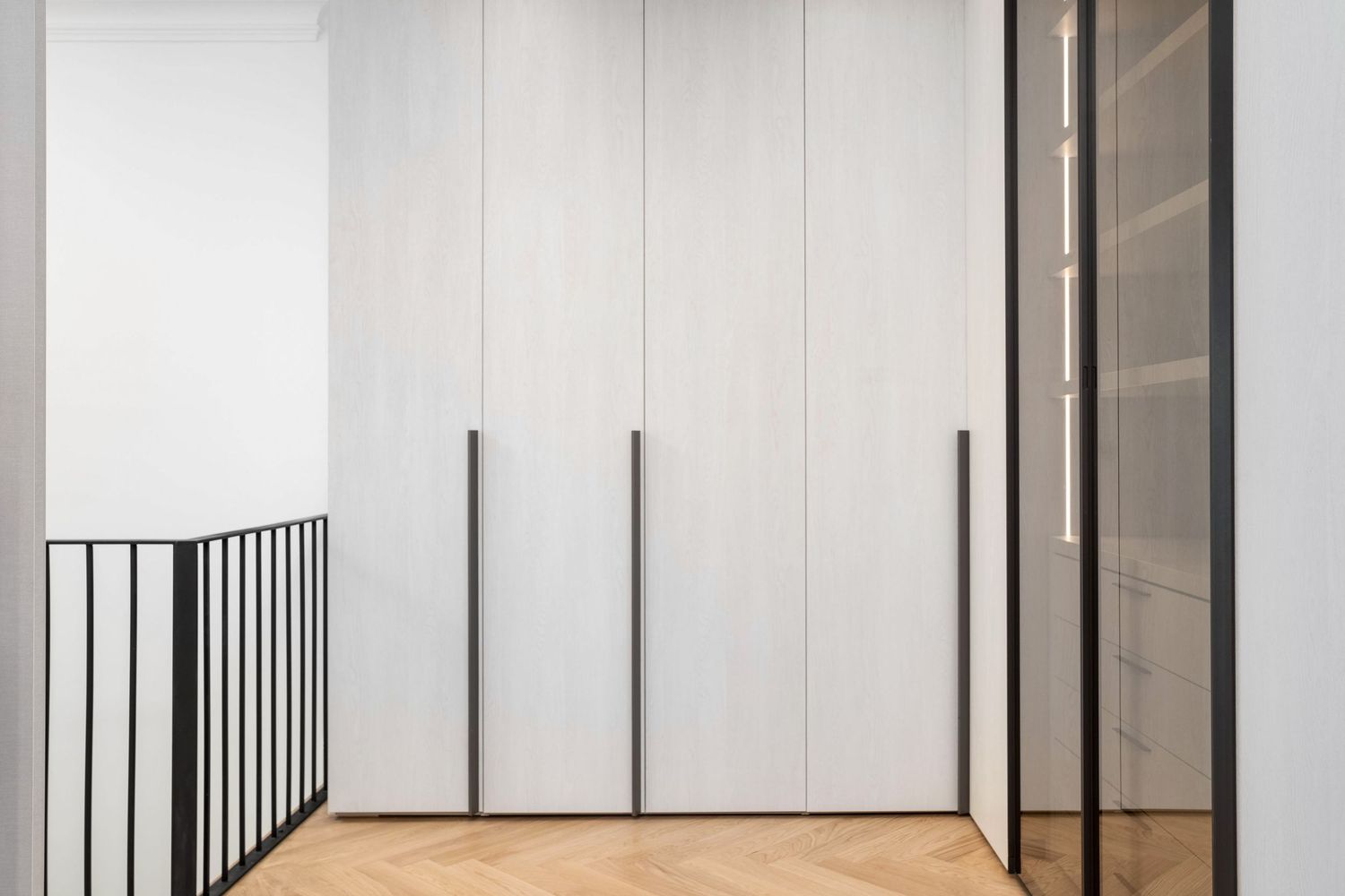 Modern hallway with floor-to-ceiling white wardrobe, black handles, herringbone parquet floor, and glass partition door.