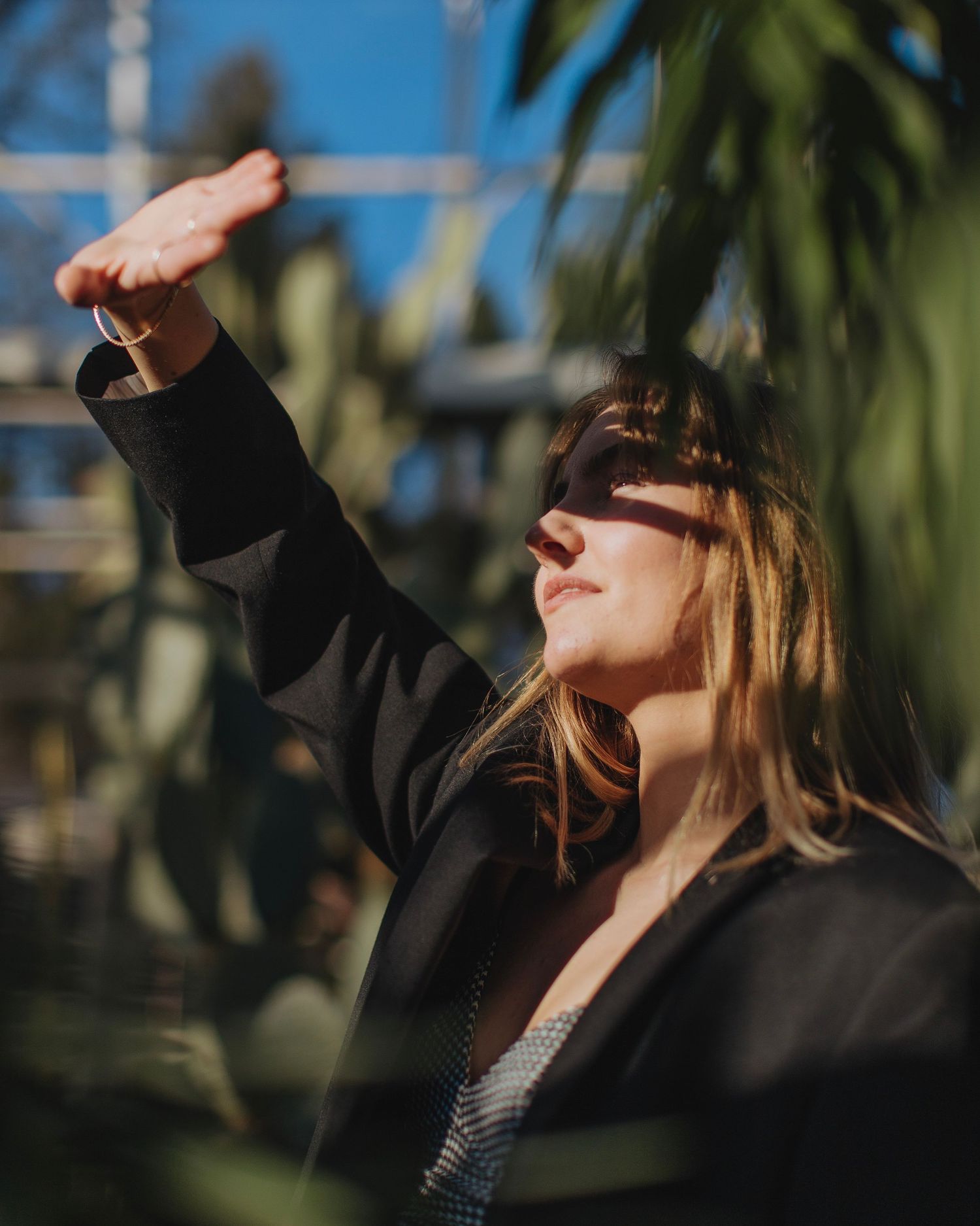 Woman in black jacket shielding eyes from bright sunlight surrounded by lush tropical greenhouse plants.
