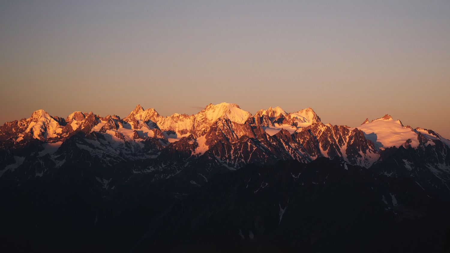 Snow-capped mountain range glowing in warm golden sunset light against a clear sky.