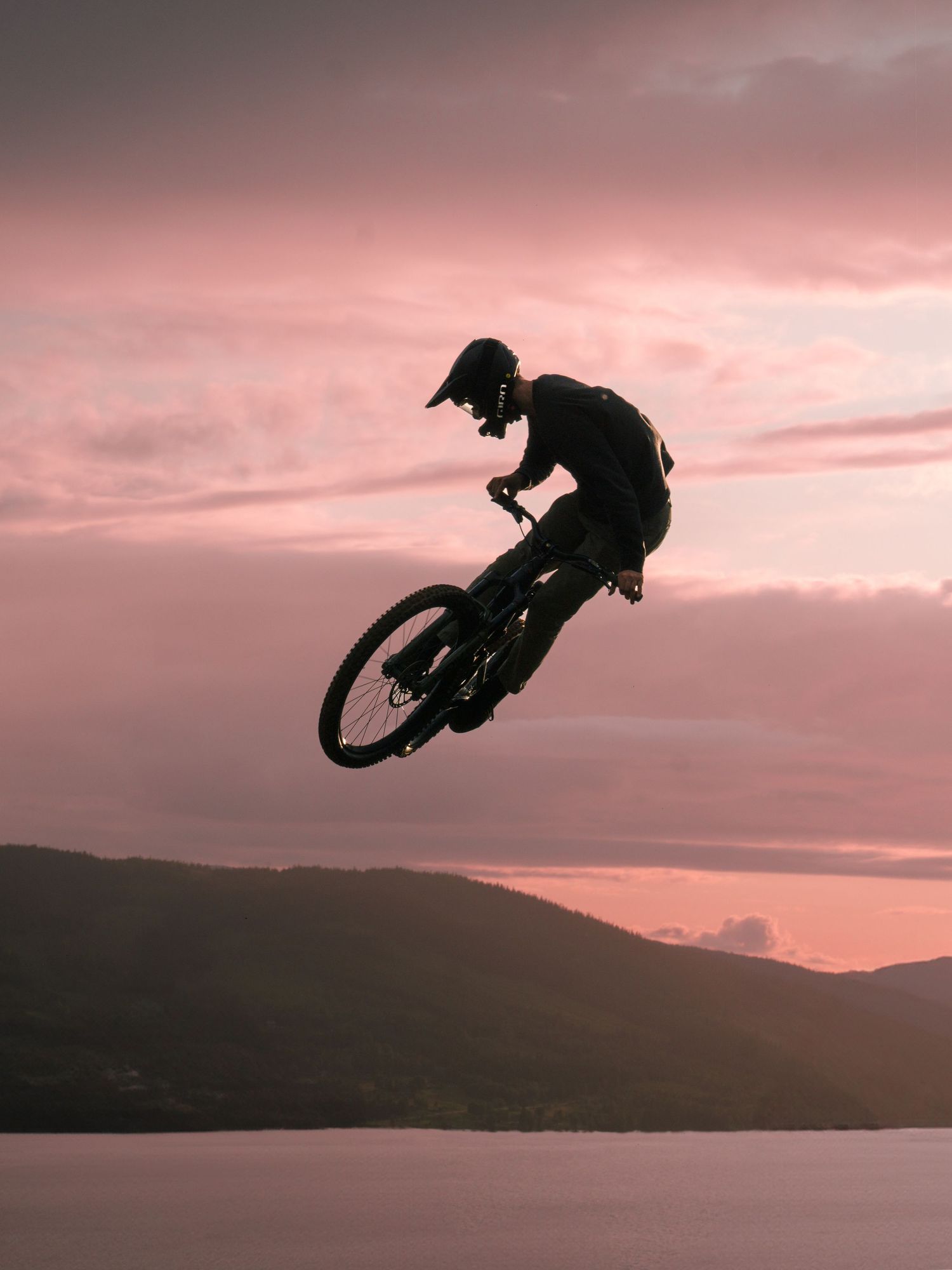 Silhouette of a mountain biker performing a jump trick against a dramatic pink sunset sky over a lake.