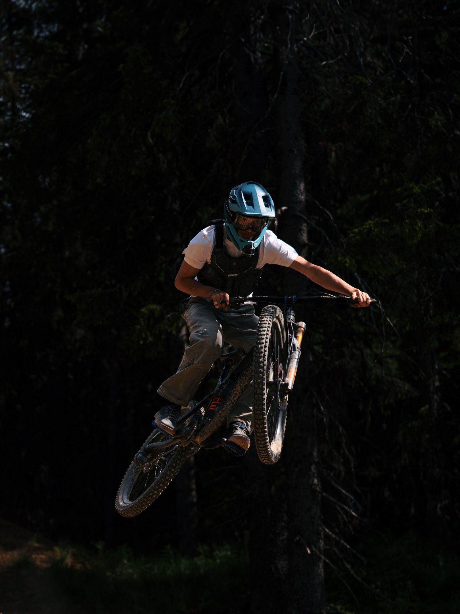 A mountain biker performs an aerial trick jump wearing a teal helmet and white shirt against a dark forest backdrop.