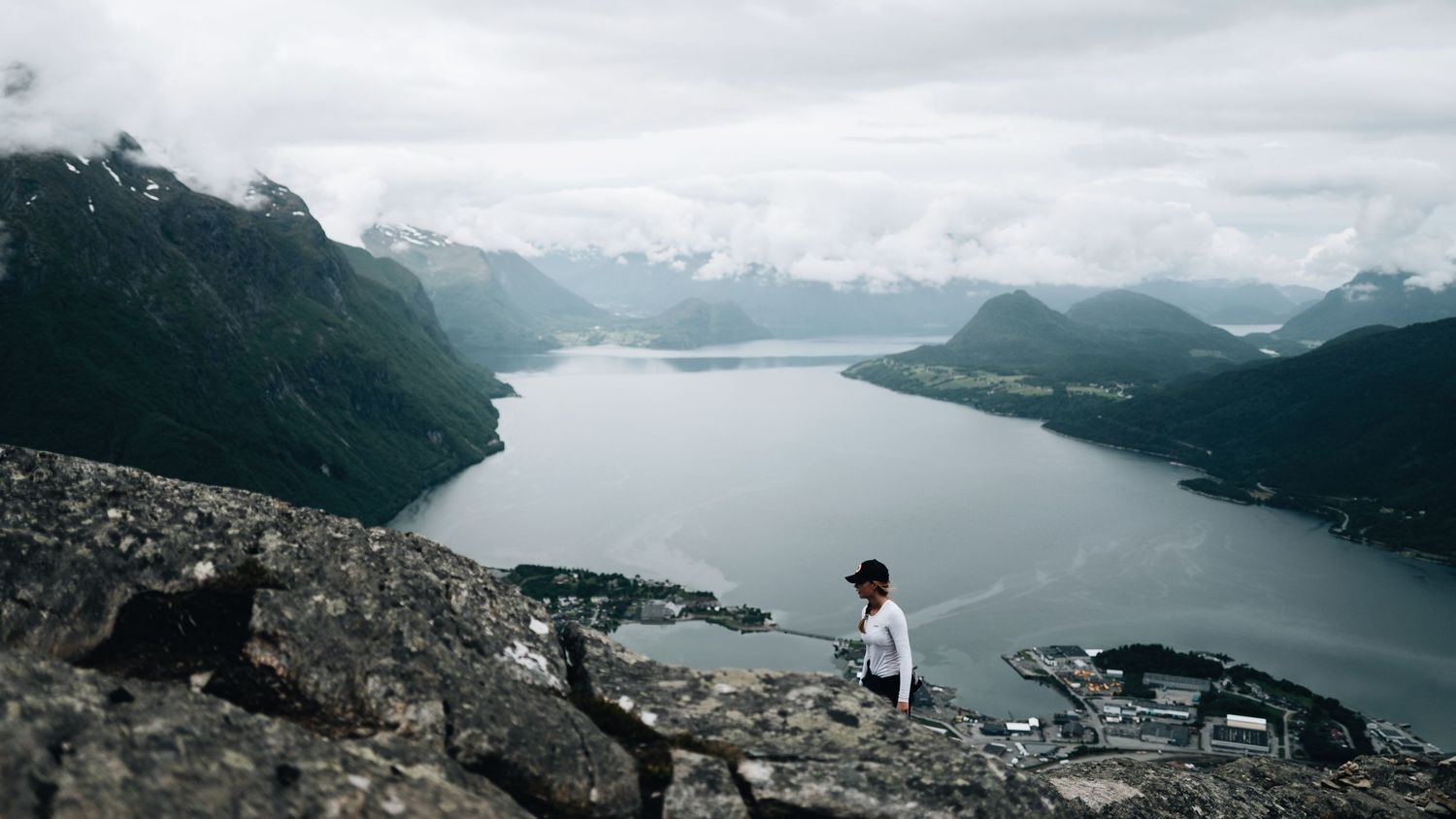A hiker stands on rocky cliffs overlooking a stunning Norwegian fjord surrounded by misty mountains and a small coastal town below.