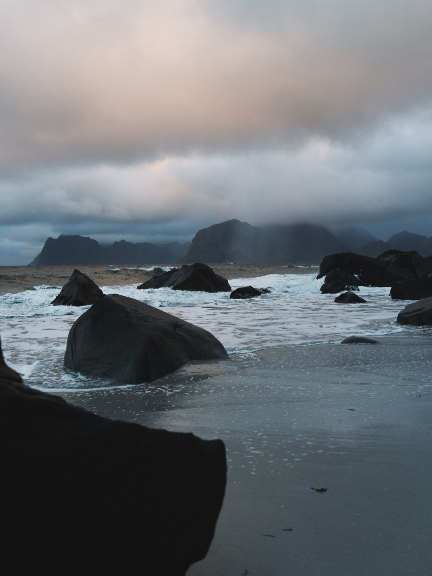 Rocky shoreline with dark boulders, crashing waves, and dramatic cloudy sky over distant mountains at dusk.