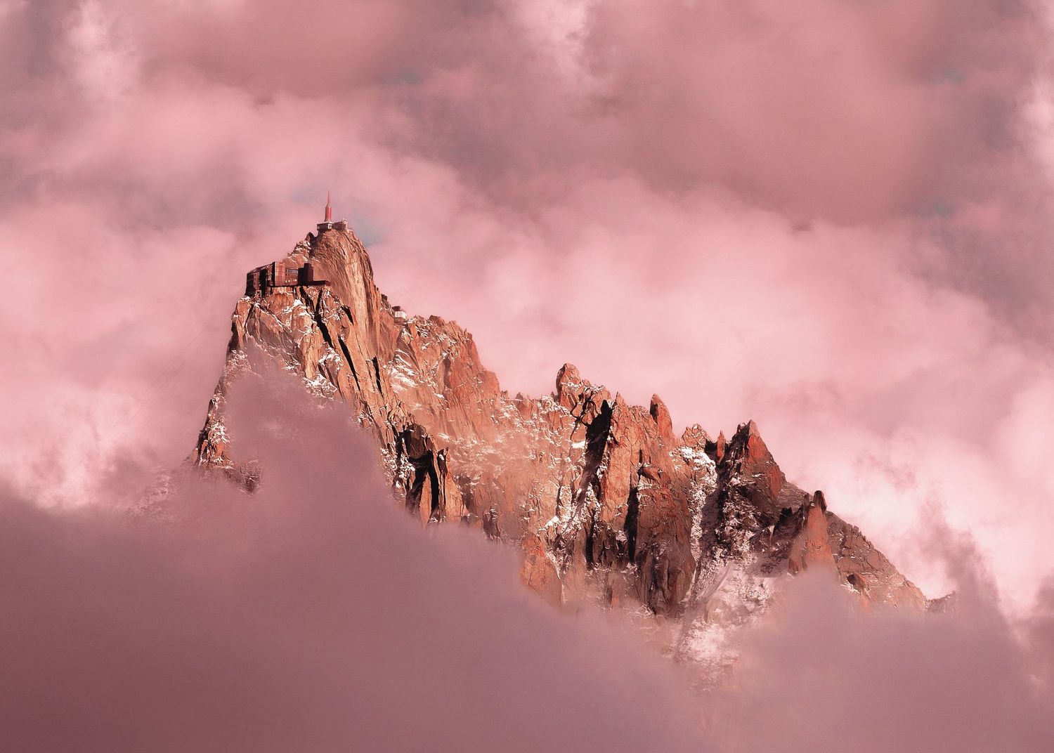 Rocky mountain peak emerging through pink-hued clouds at sunset, with jagged stone spires against a soft rose sky.
