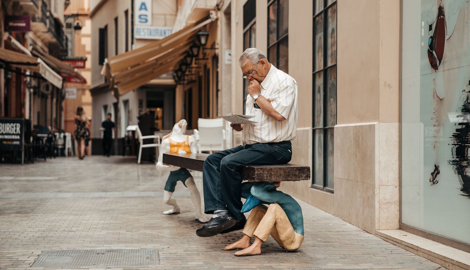 Elderly man sitting on a bench in a European pedestrian street, reading while enjoying a snack outdoors.