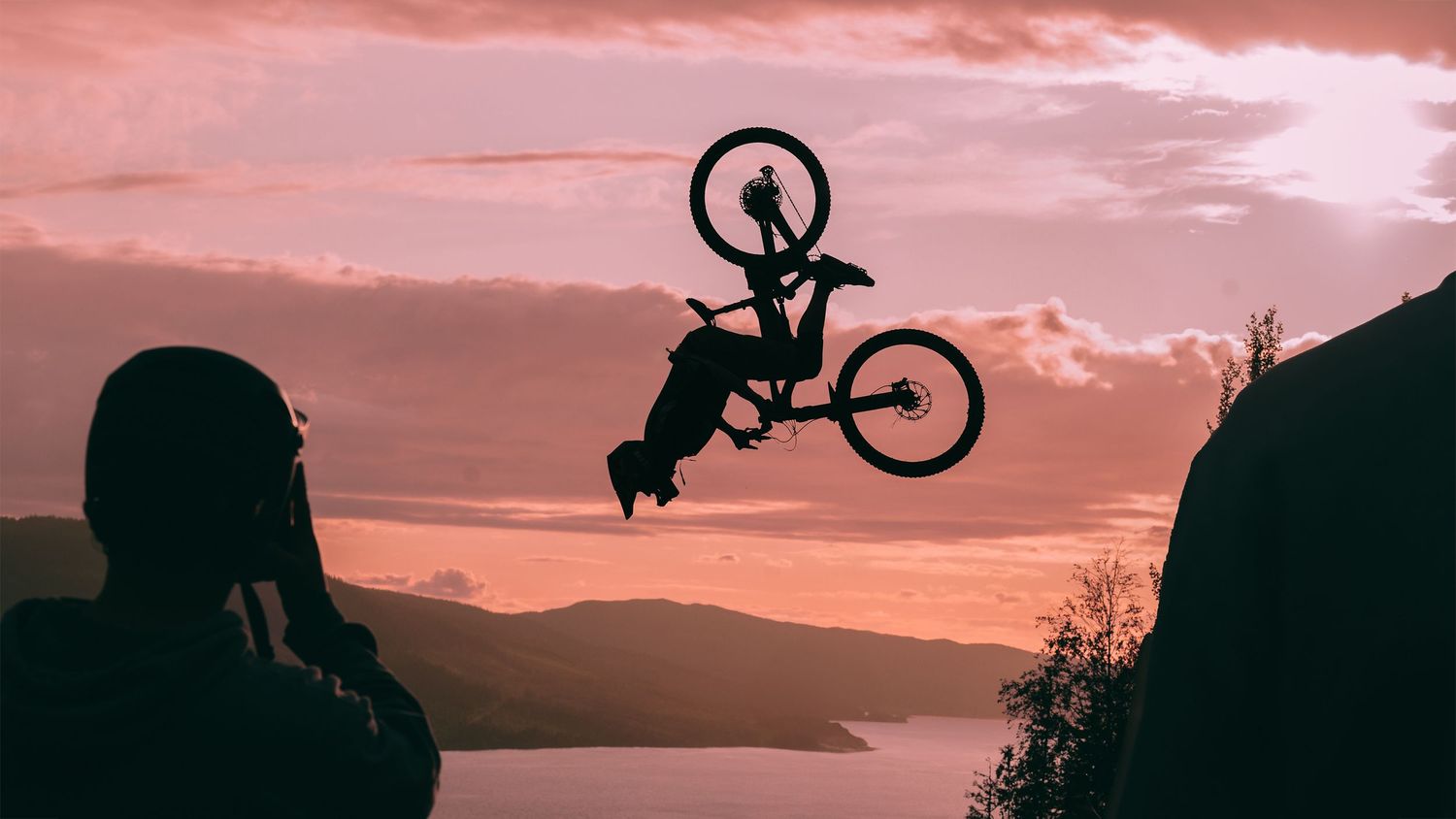 Silhouette of a BMX rider performing a dramatic aerial trick at sunset over a scenic lake landscape.