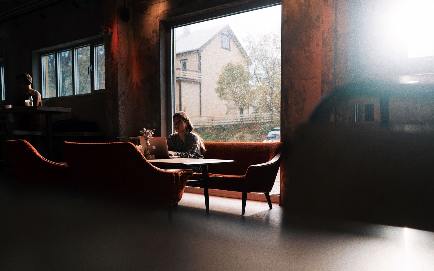 Person sitting alone at a table in a moody cafe with large windows and warm leather seating.