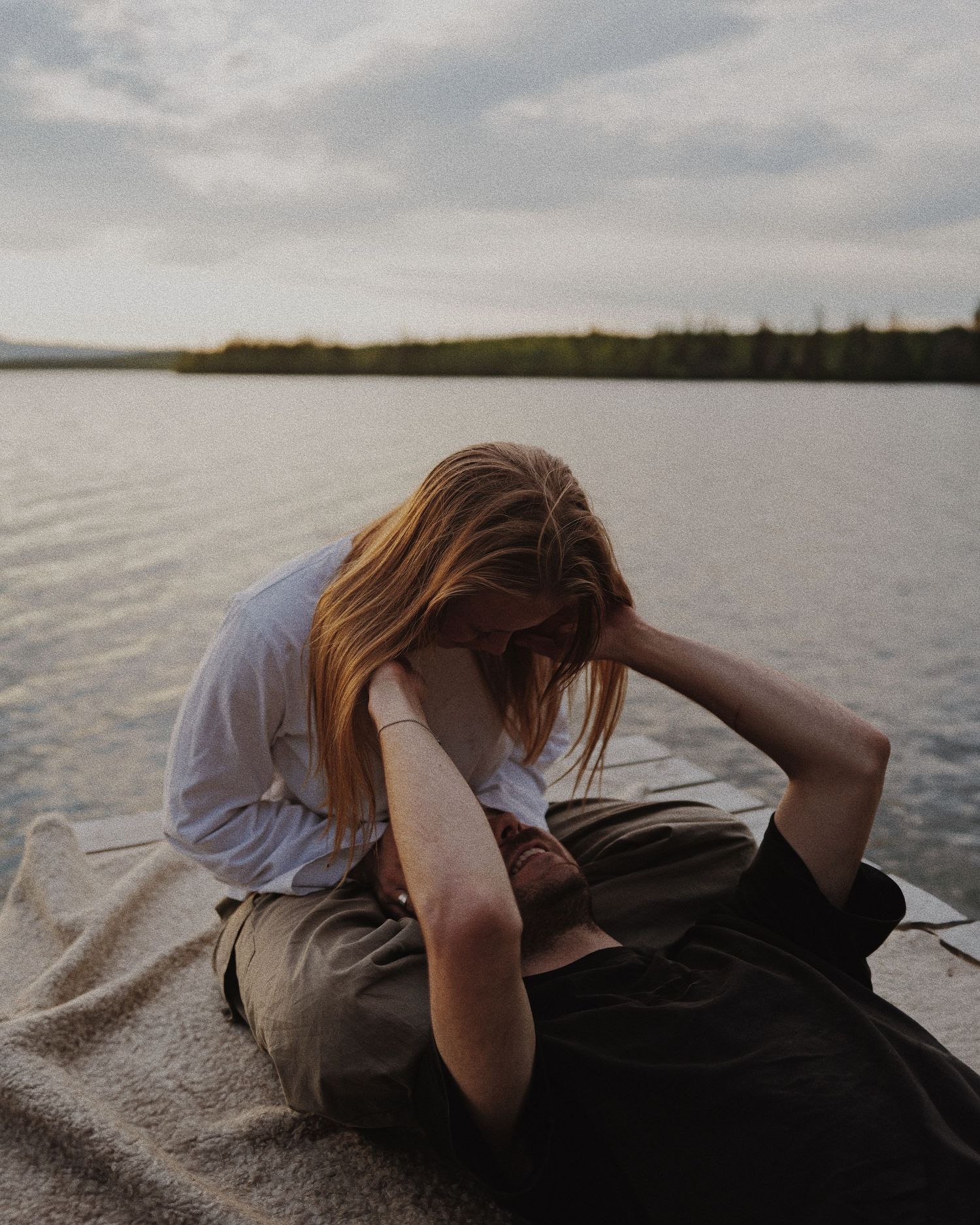 Young woman sitting alone on a dock by a calm lake, head down in a contemplative pose at dusk.