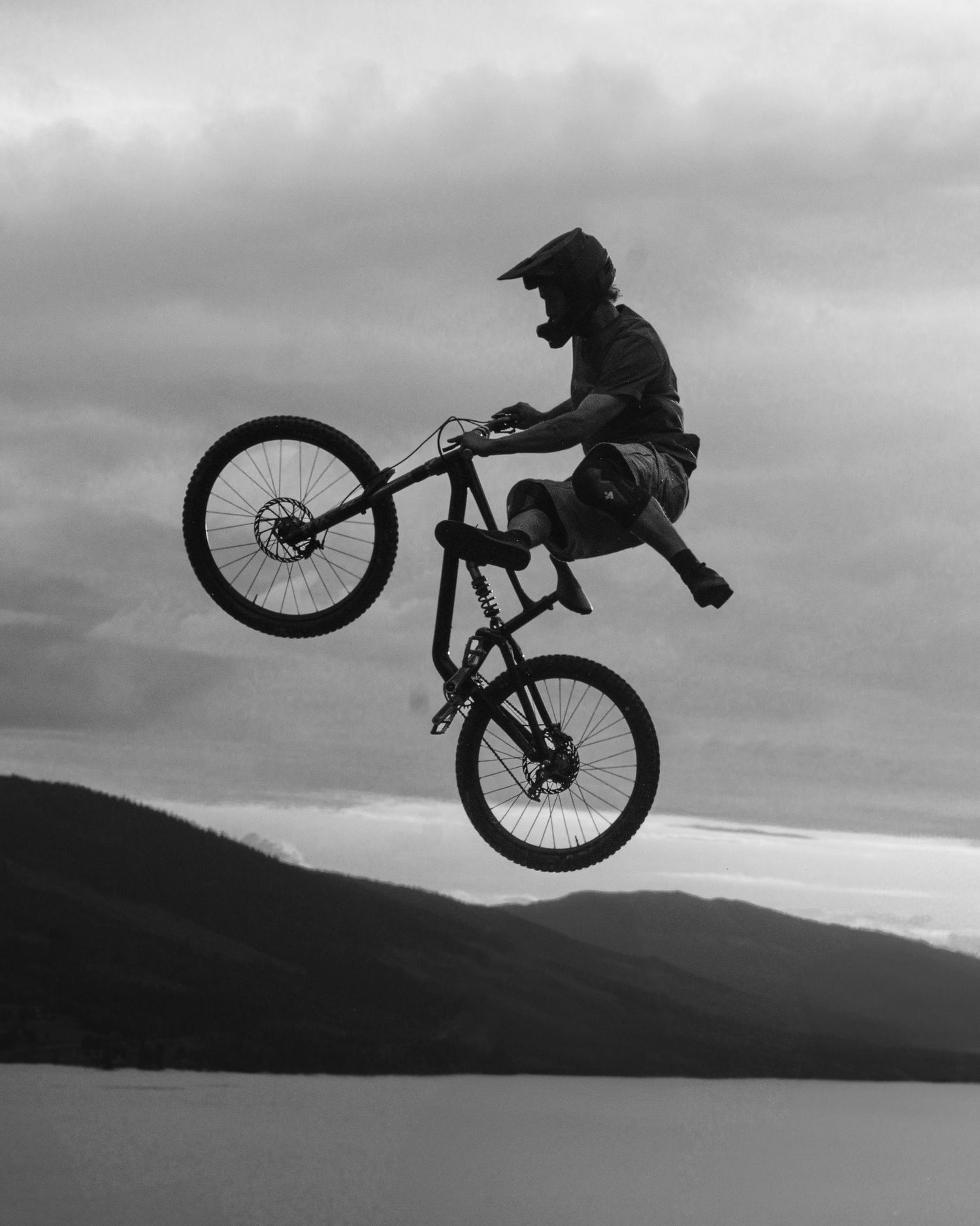 A daring mountain biker performs a high-flying aerial trick against a dramatic cloudy sky with mountain lake backdrop.