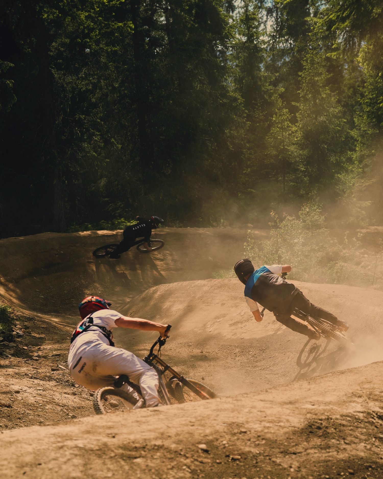 Three mountain bikers race through a dusty forest trail, kicking up dirt clouds on a sunny day.