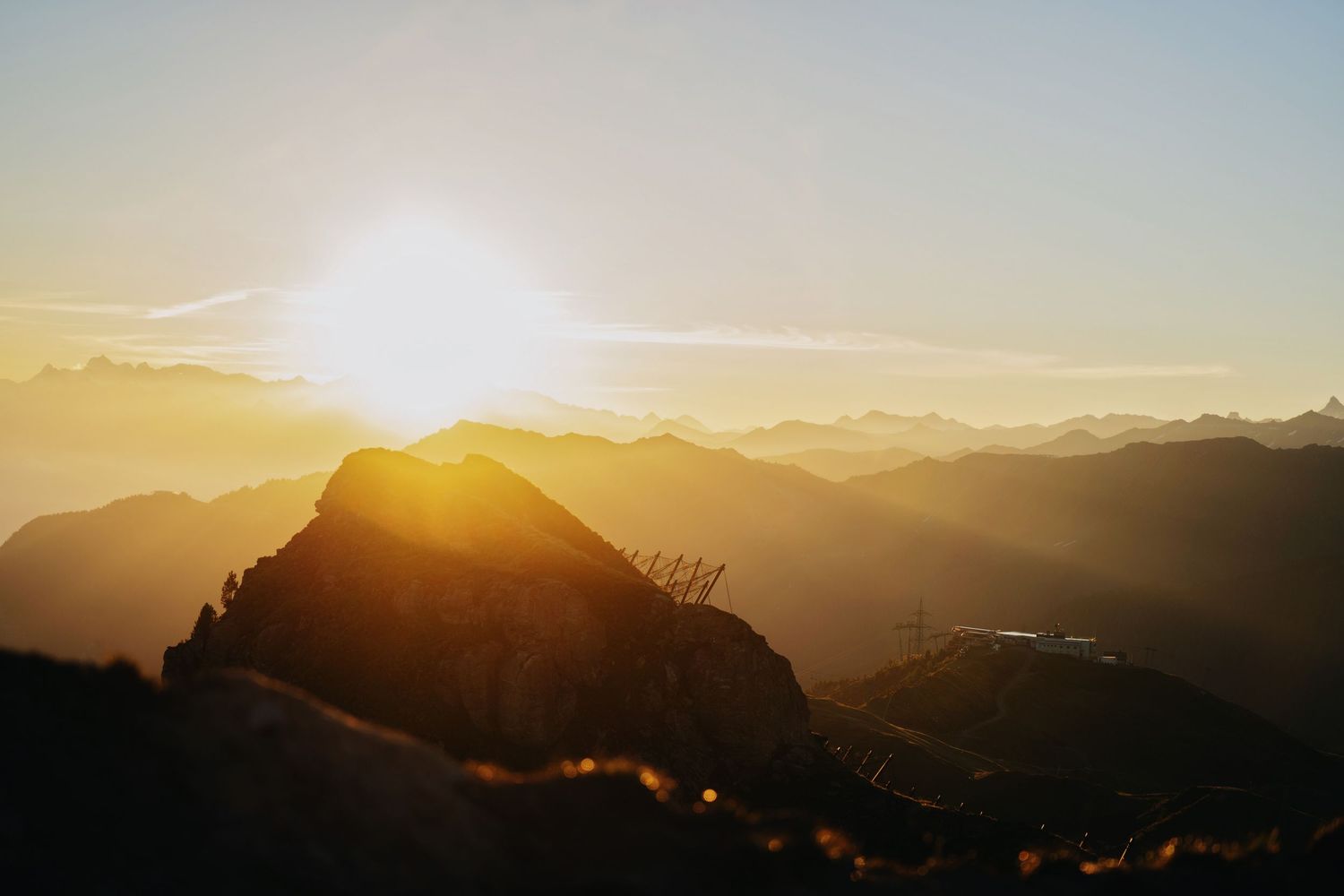Golden sunset behind a mountain peak with layered silhouettes of Alpine ranges glowing in warm orange light.