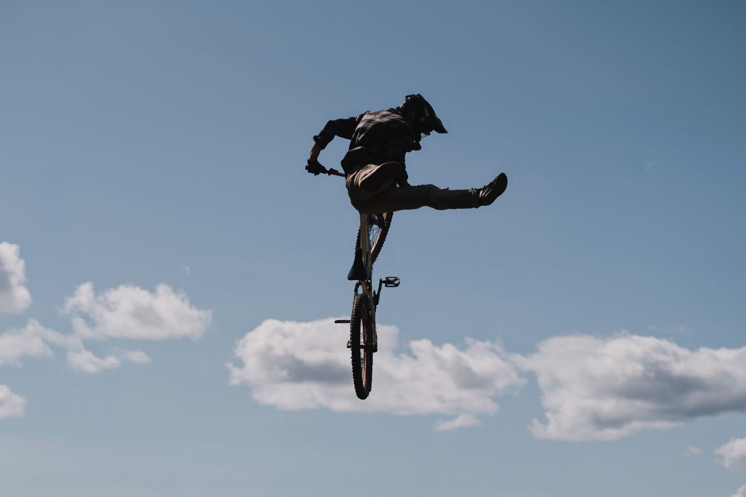 BMX rider performs a dramatic aerial trick against a blue sky with scattered clouds, silhouetted mid-air.