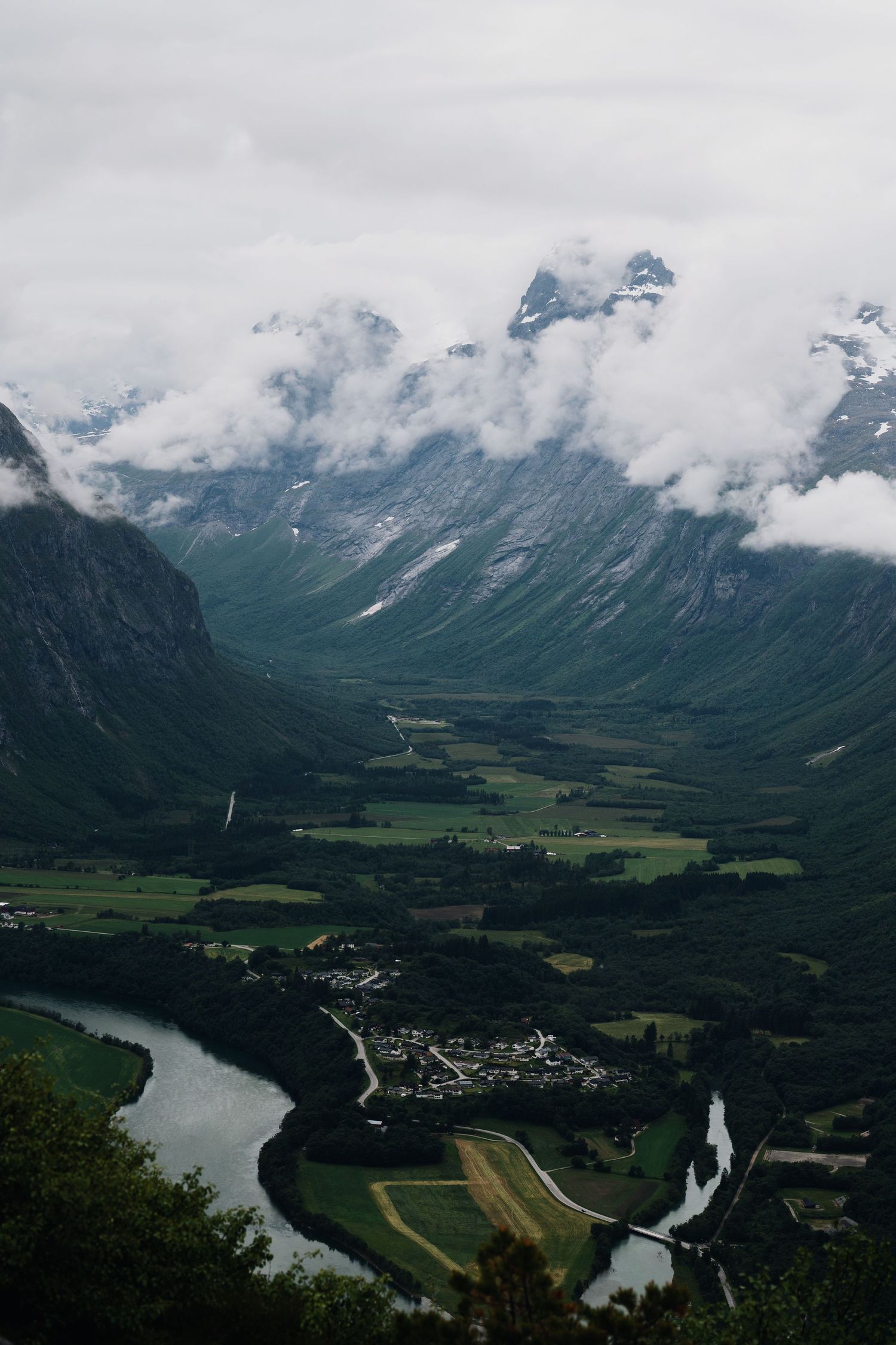 Aerial view of a lush green Norwegian valley with a winding river, small town, and cloud-covered mountains.