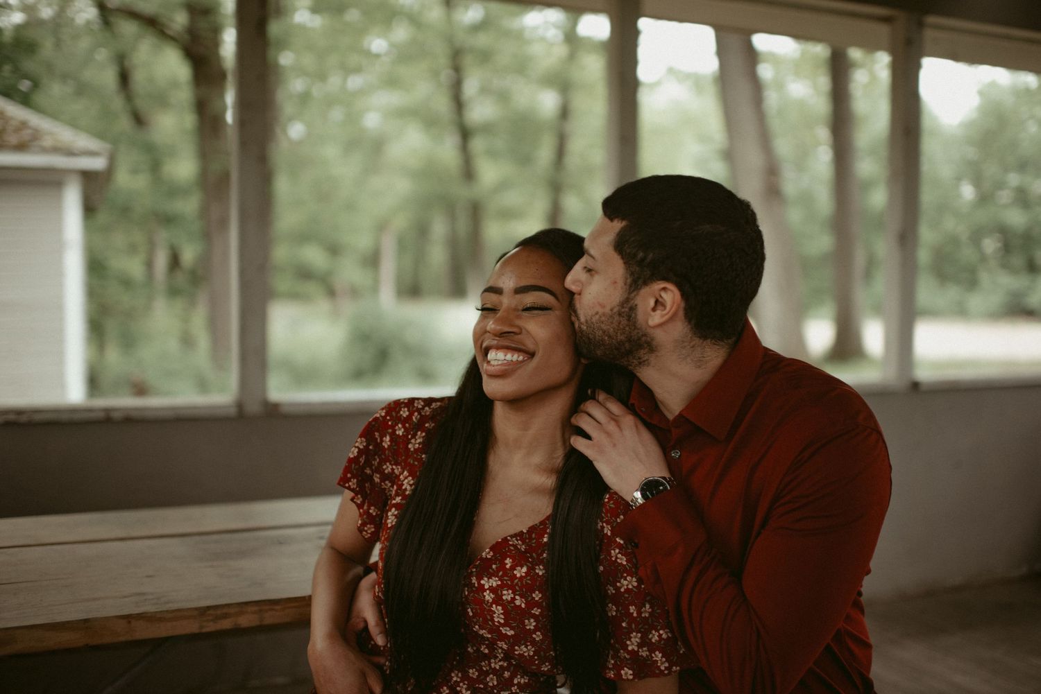Happy couple sharing a tender kiss moment in an outdoor pavilion, woman laughing joyfully in red floral dress.