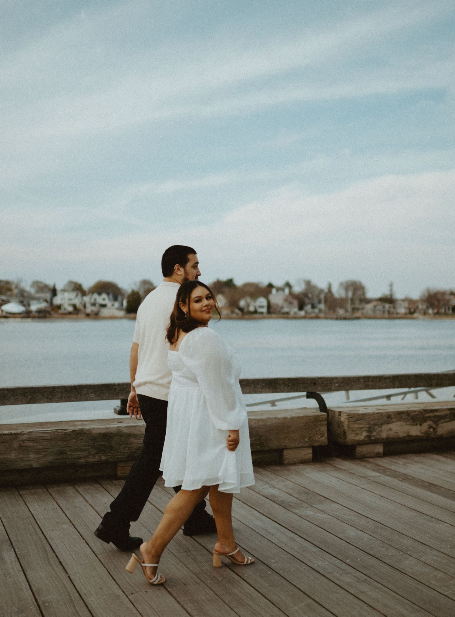 Smiling woman in white dress walking with partner along a waterfront boardwalk on a cloudy day.
