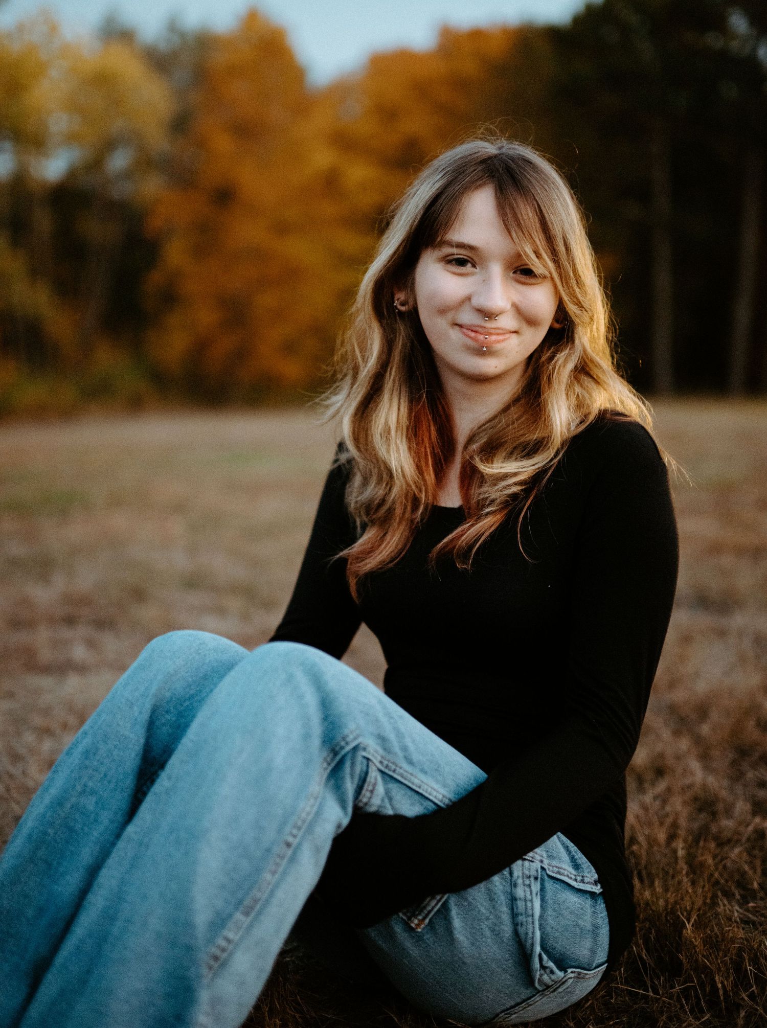 Young woman with wavy hair sitting in a field wearing jeans and black top surrounded by golden autumn trees.