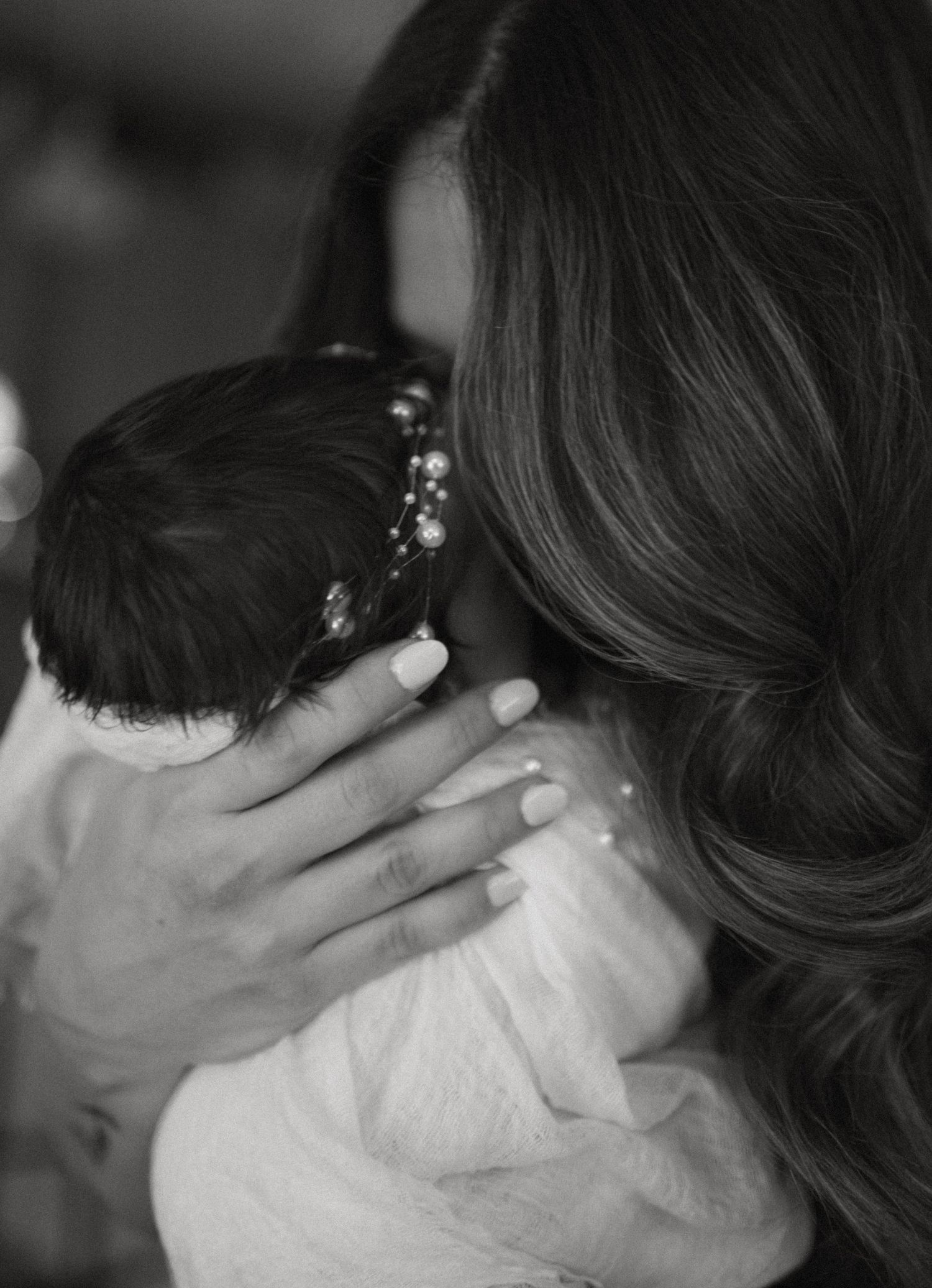 Black and white photo of mother kissing newborn baby with pearl headband, showing tender bond.