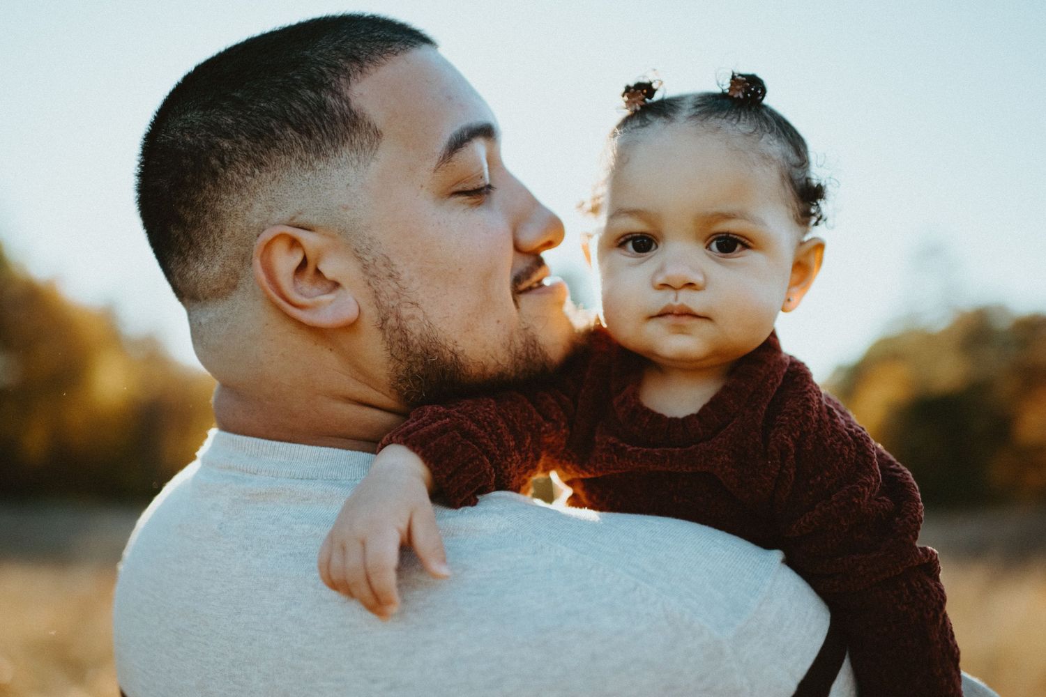 Father holding toddler daughter close in warm golden sunset light outdoors.
