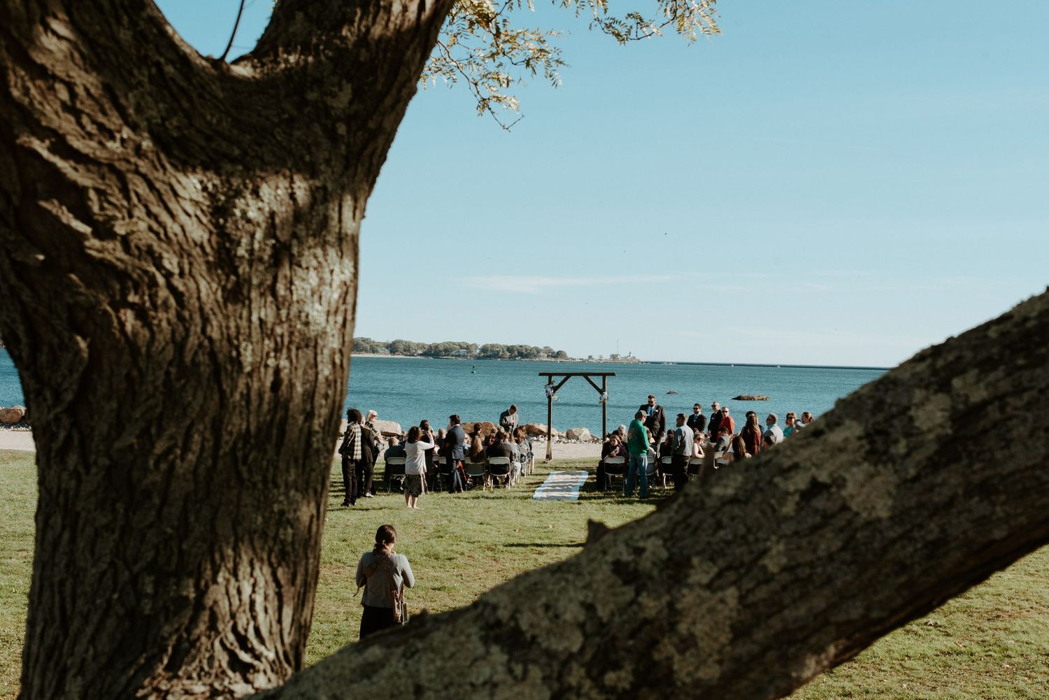Outdoor waterfront wedding ceremony framed by large tree trunks with guests seated on green lawn.
