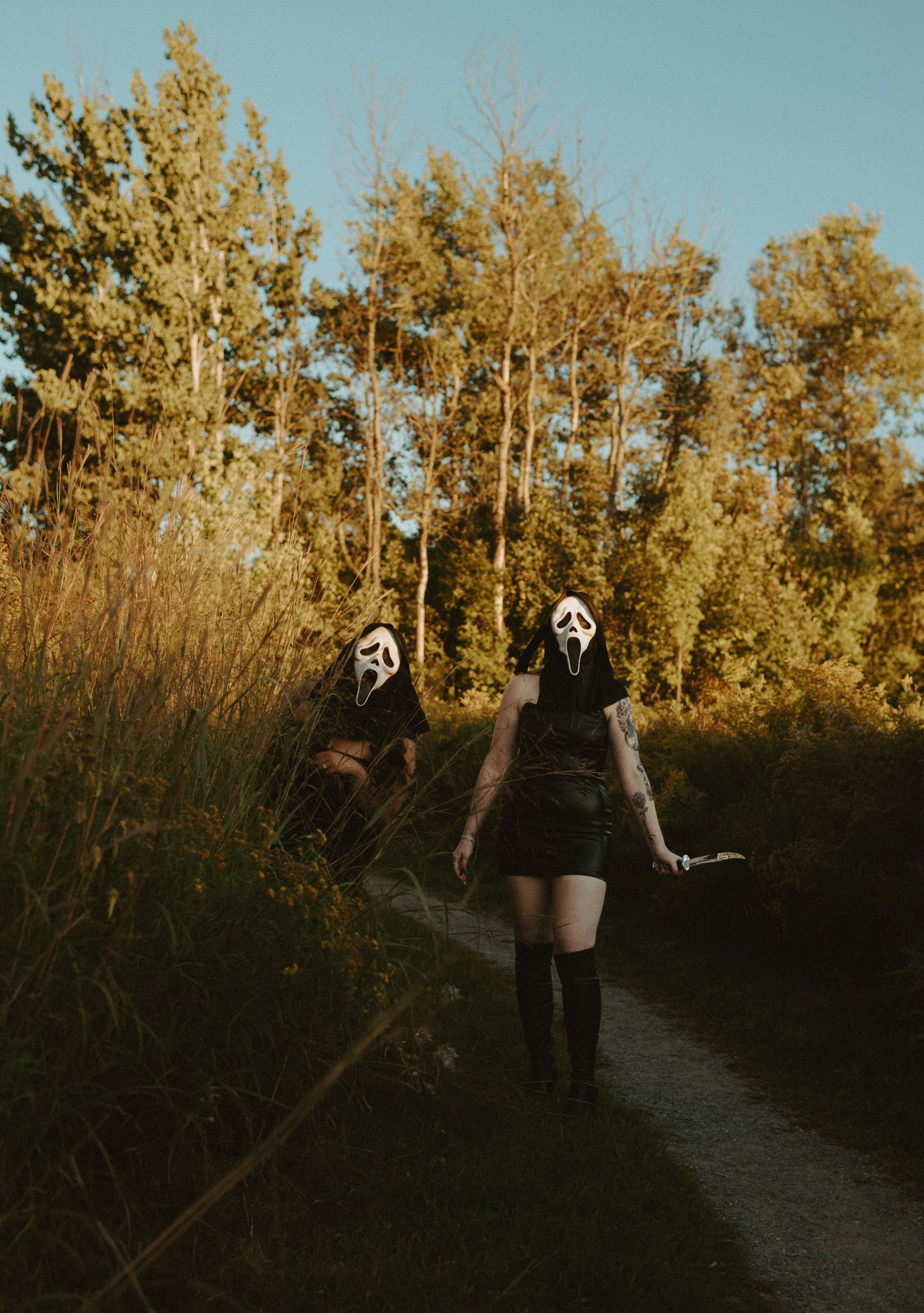 Two people wearing Ghostface masks and black robes walking on a nature trail in Halloween costumes.
