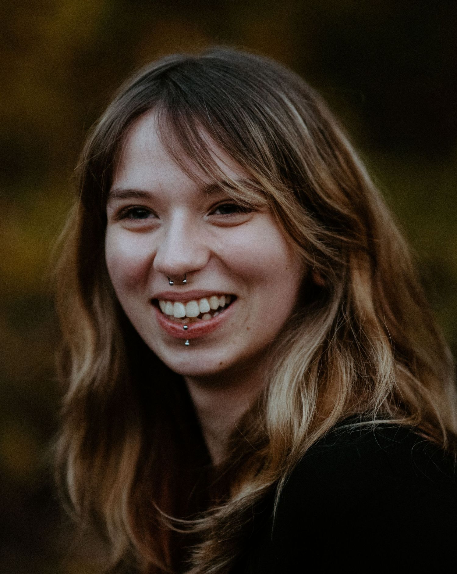 Smiling young woman with wavy hair laughing outdoors against blurred autumn foliage.