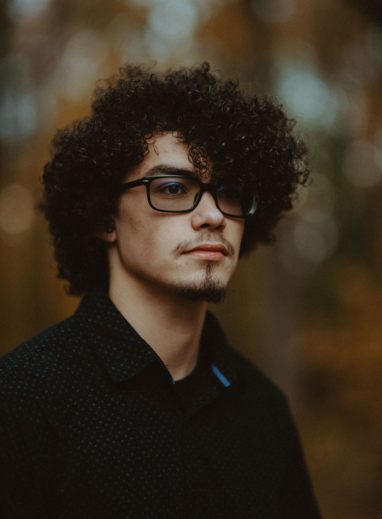 Young man with curly hair and glasses posing in autumn bokeh background.