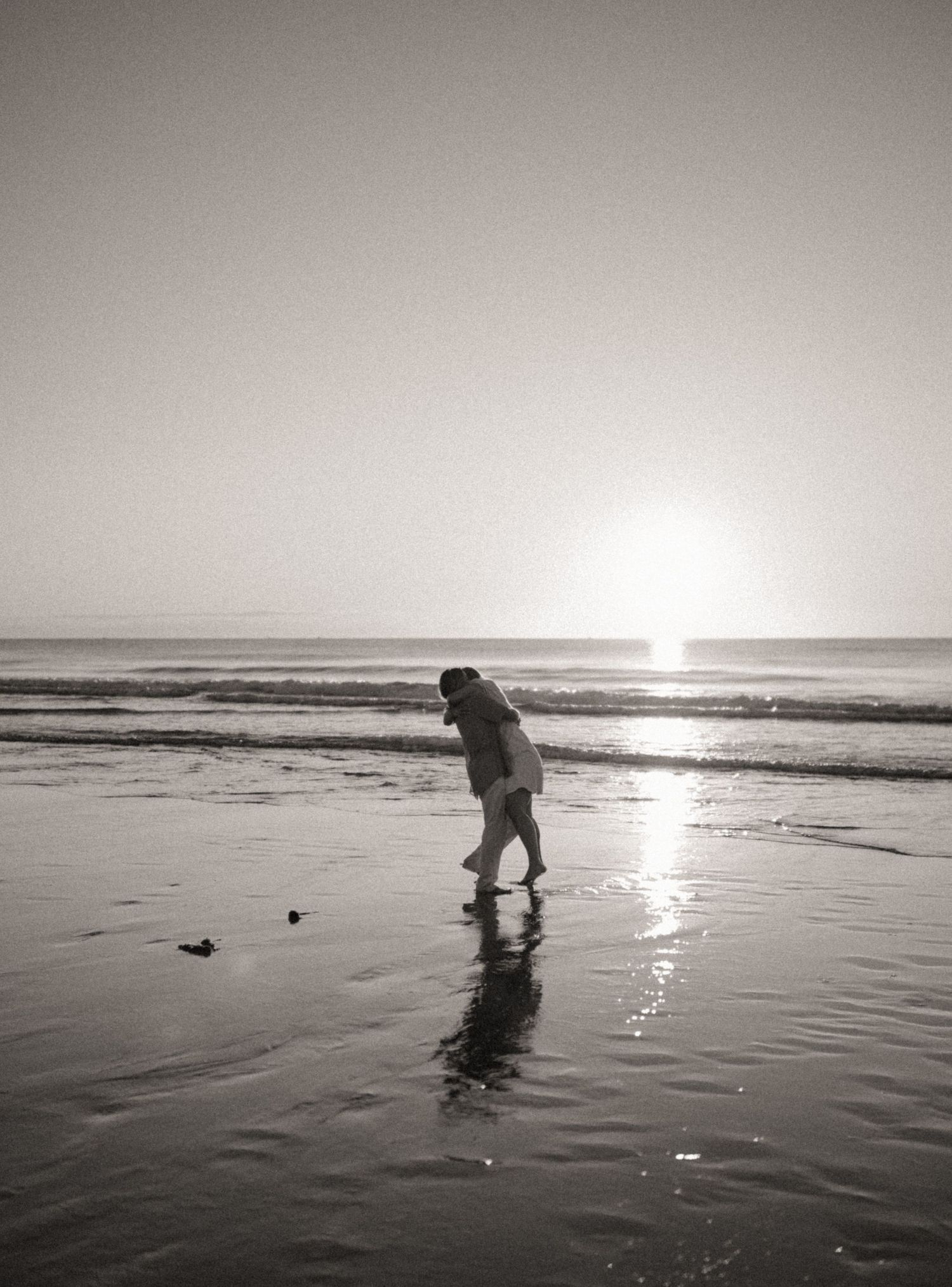Black and white photo of couple embracing on reflective beach at sunrise by ocean waves.