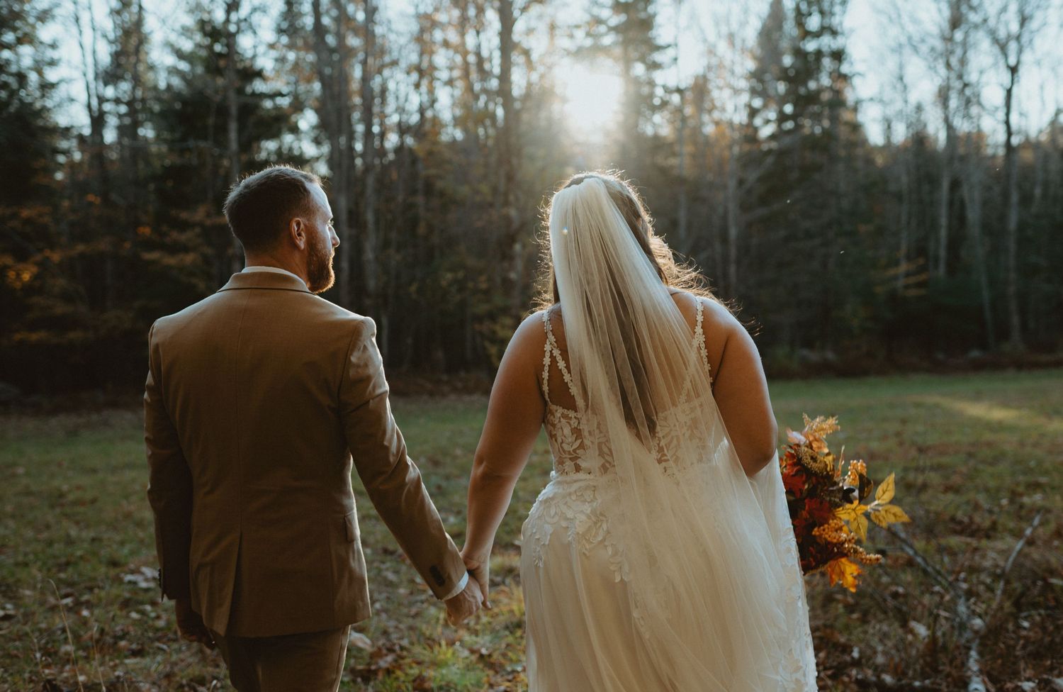 Bride and groom holding hands facing a sunlit forest in an outdoor autumn wedding setting.