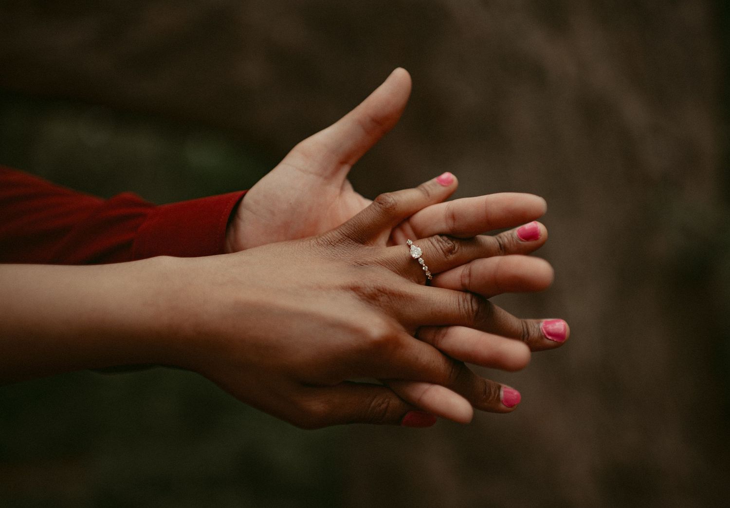 Close-up of intertwined hands showing an engagement ring, with pink manicured nails against a blurred background.