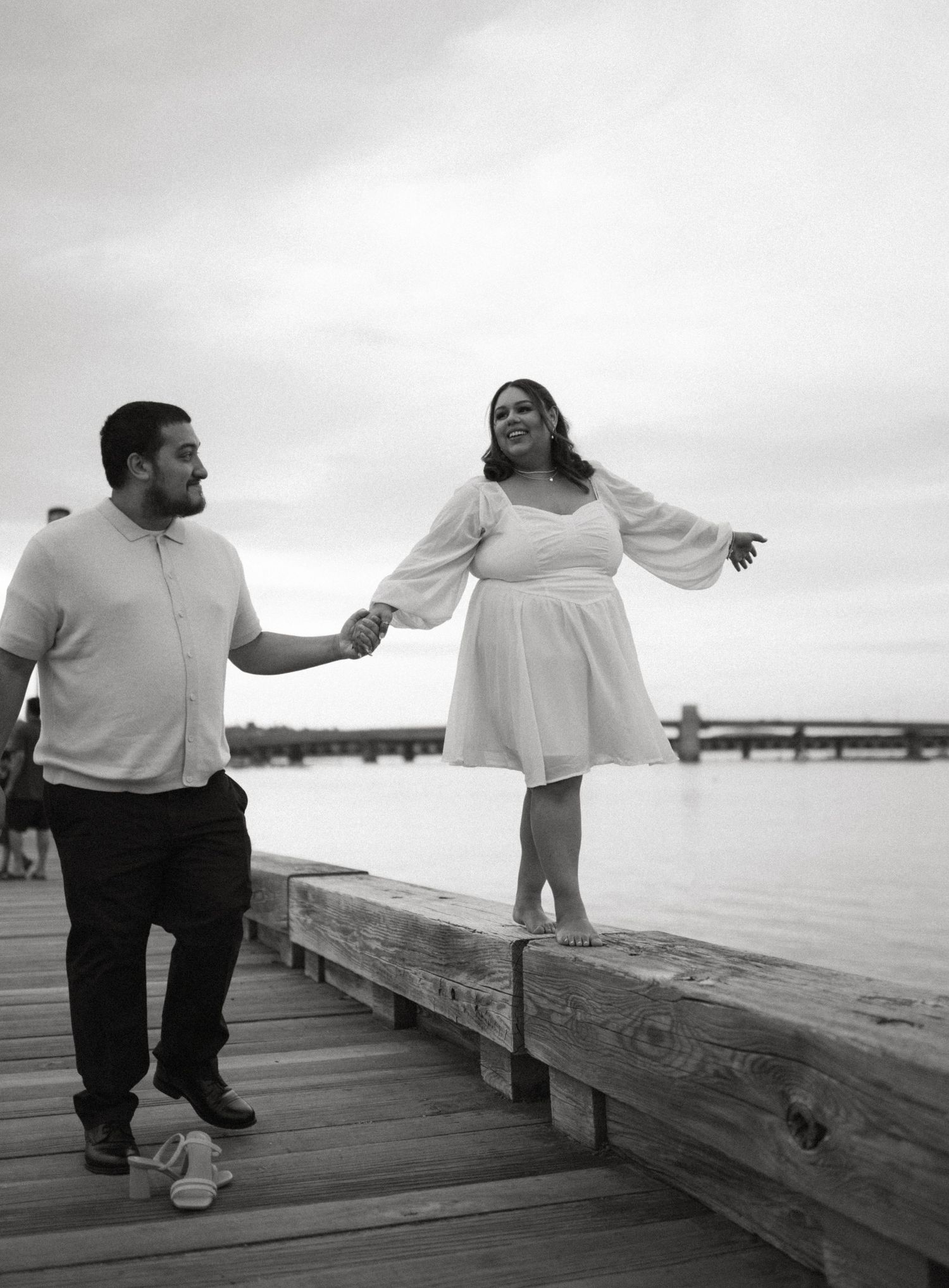 Black and white photo of a couple holding hands on a waterfront dock, woman in white dress balancing on edge.
