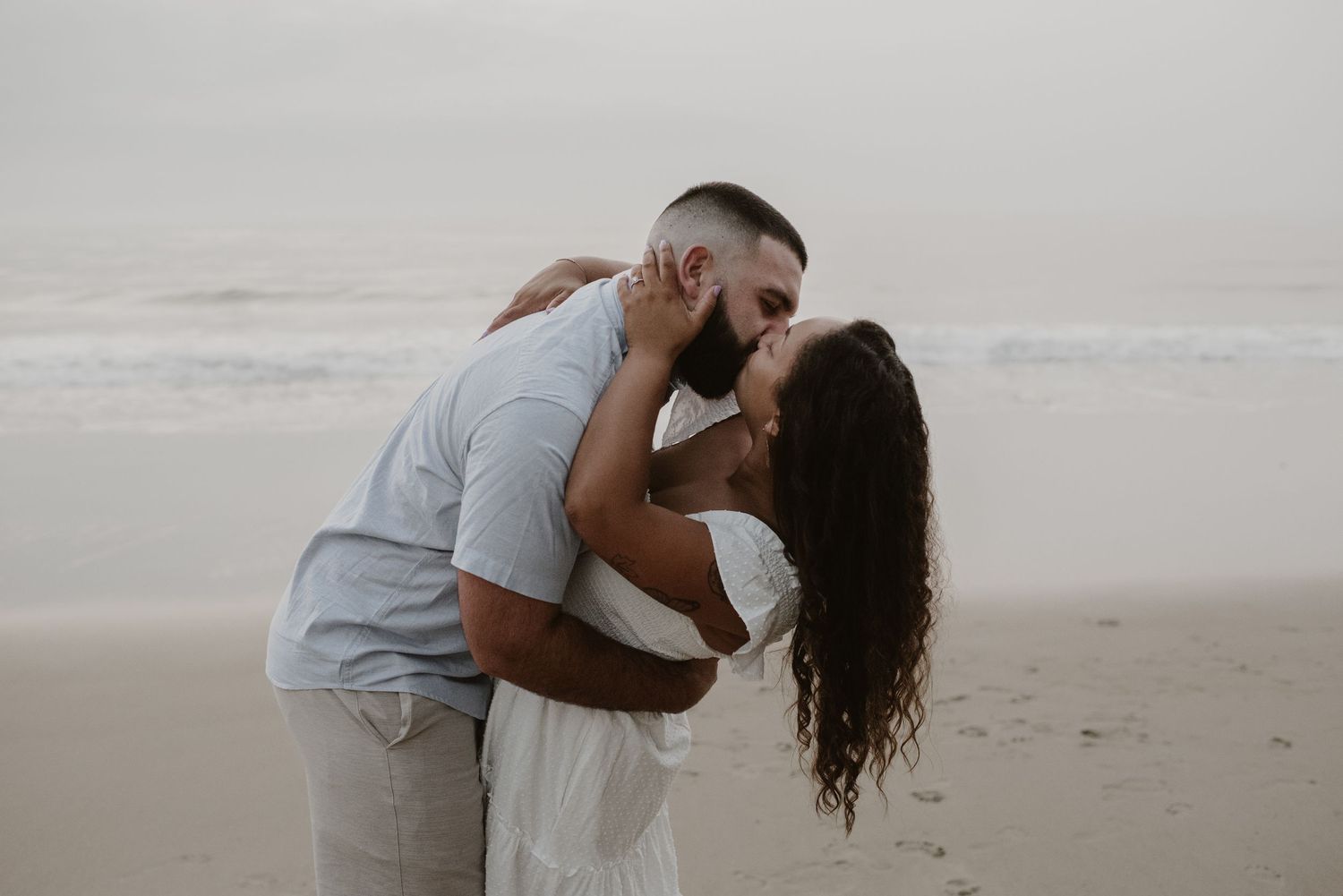 Romantic couple sharing a kiss on a foggy beach during an engagement photo session.