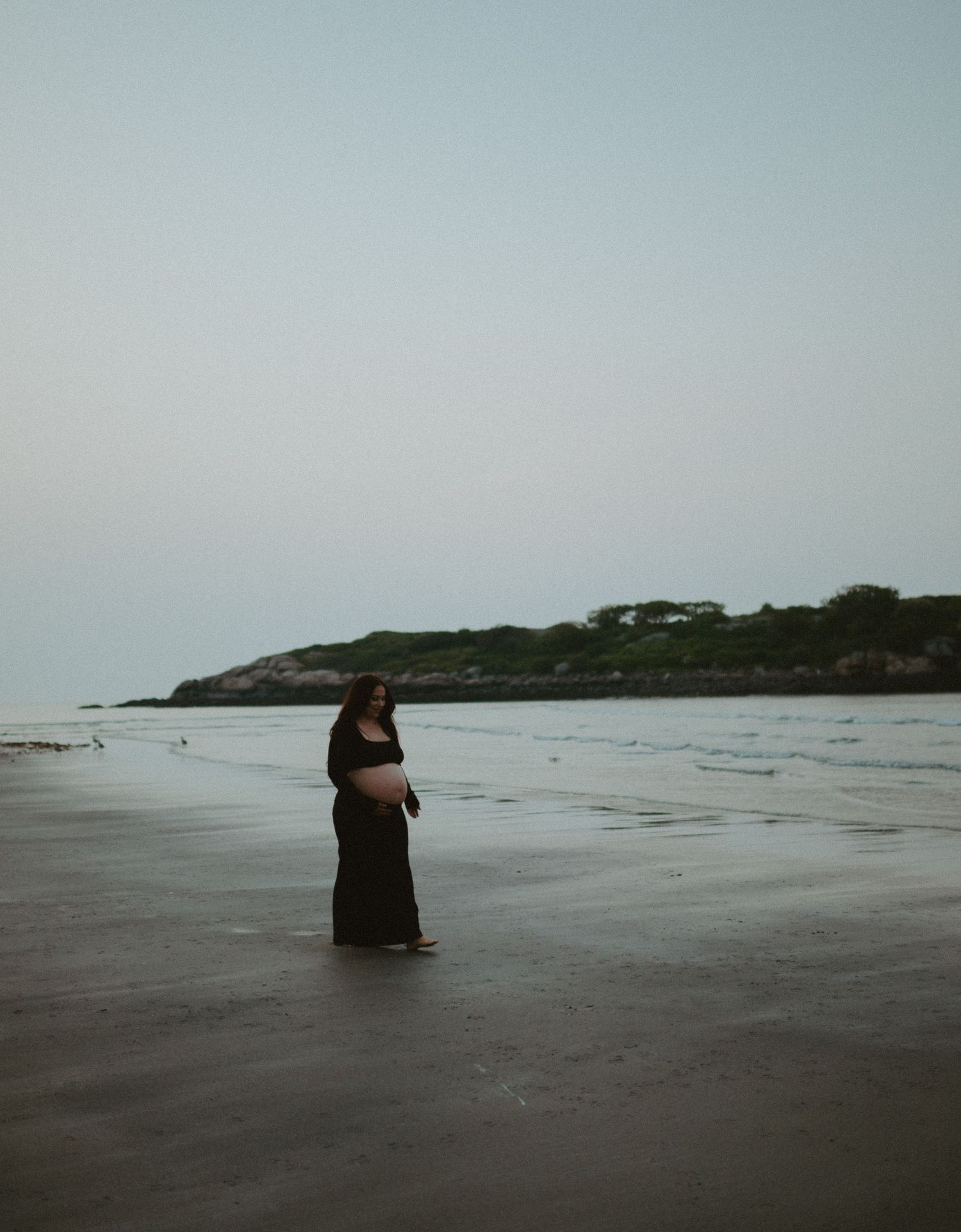 Pregnant woman in black dress stands on wet sand beach with rocky coastline in background.