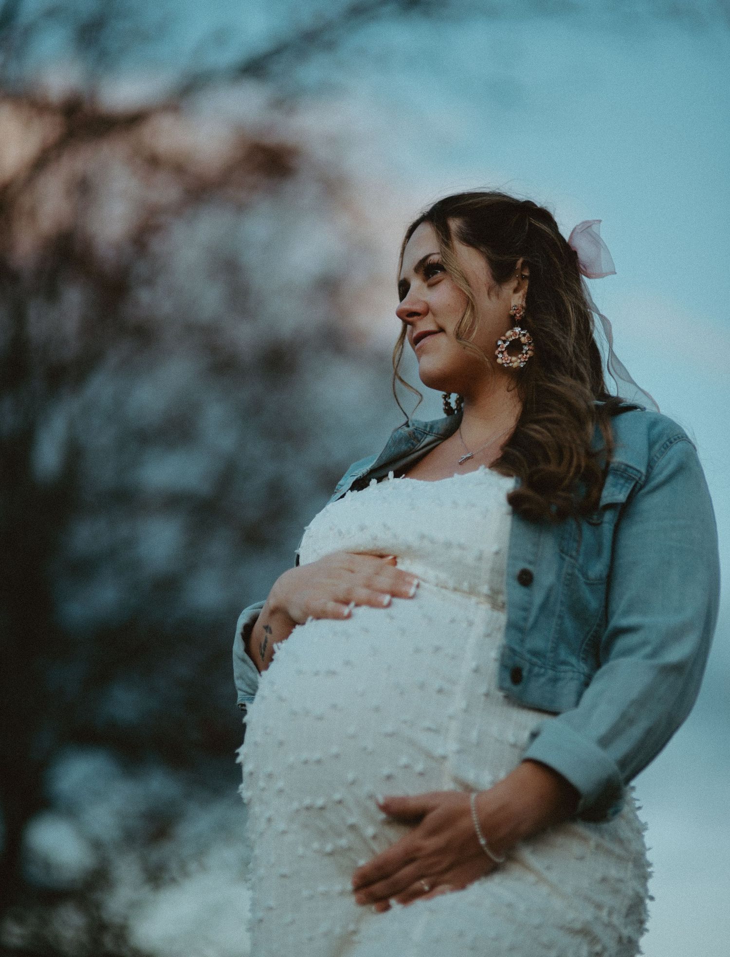 Glowing pregnant woman in white dress and denim jacket holds baby bump while smiling outdoors at sunset.