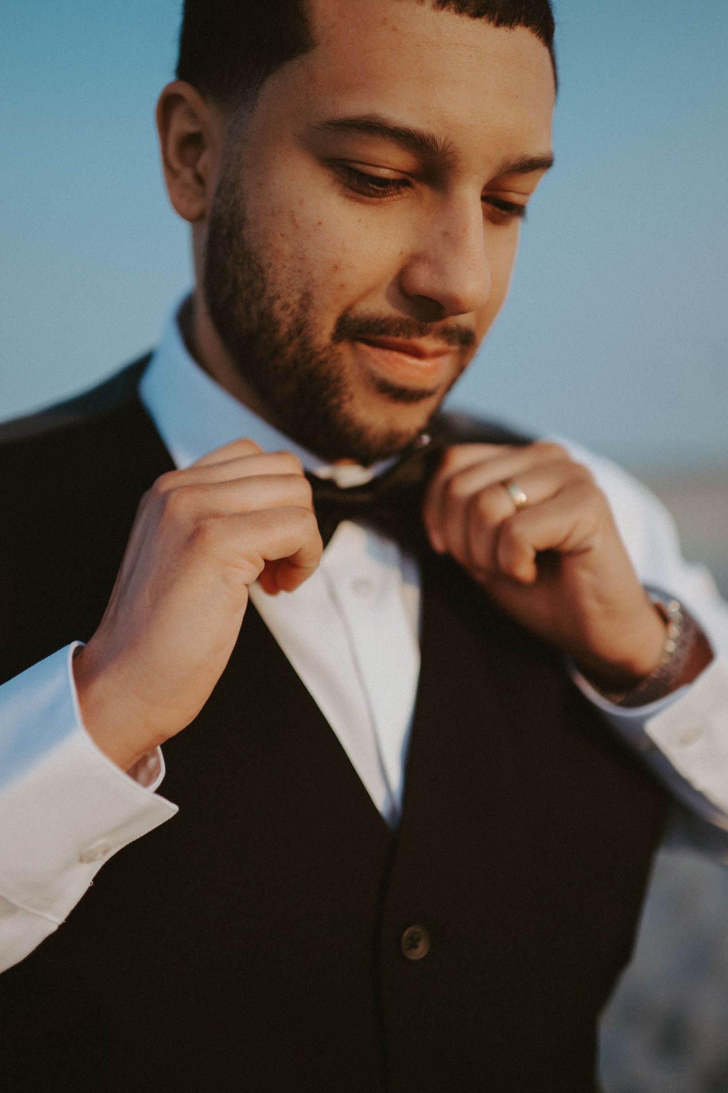 Groom adjusting black bow tie wearing white shirt and black vest with watch and ring at golden hour.