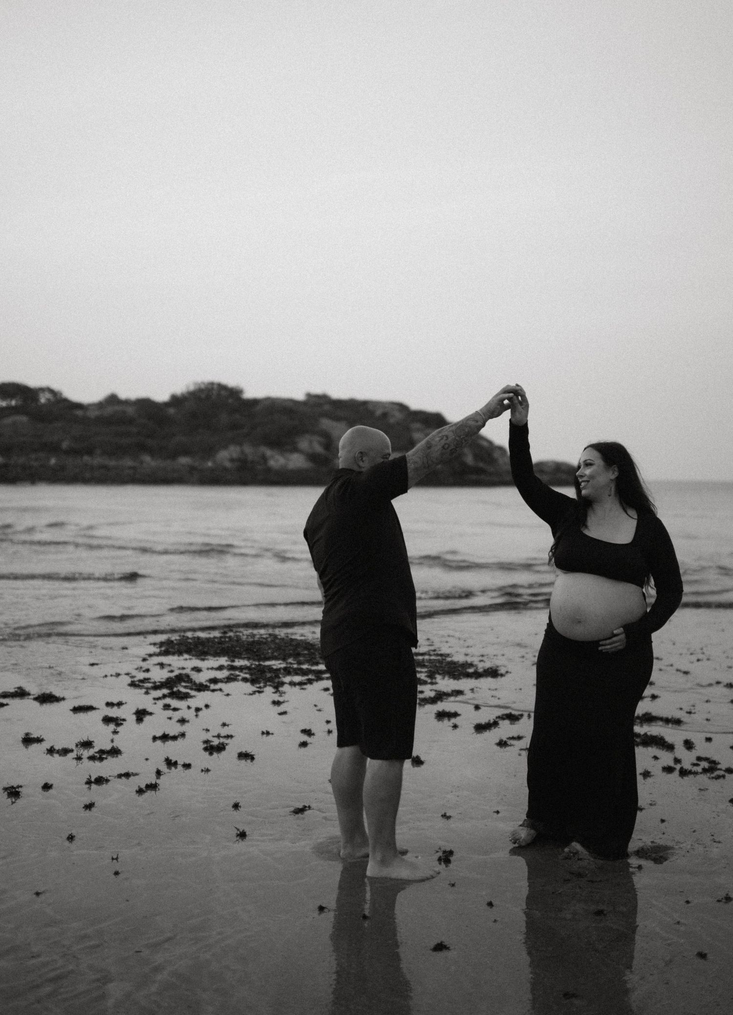 Black and white photo of couple dancing on beach, man is spinning pregnant woman near ocean.