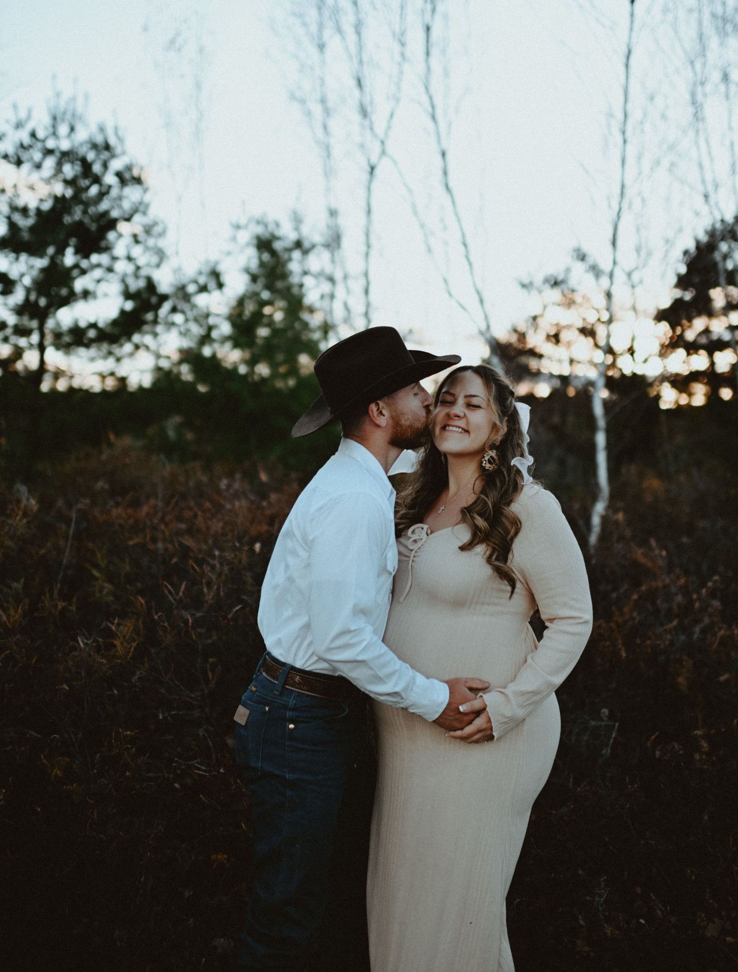 Man in black cowboy hat kisses pregnant woman in cream maternity dress during outdoor fall photo session.
