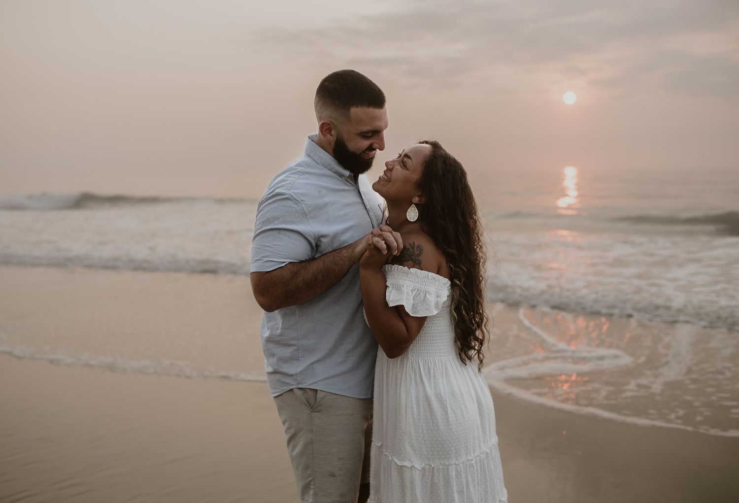 Couple embracing on sandy beach at sunset, woman in white off-shoulder dress and man in light blue shirt.