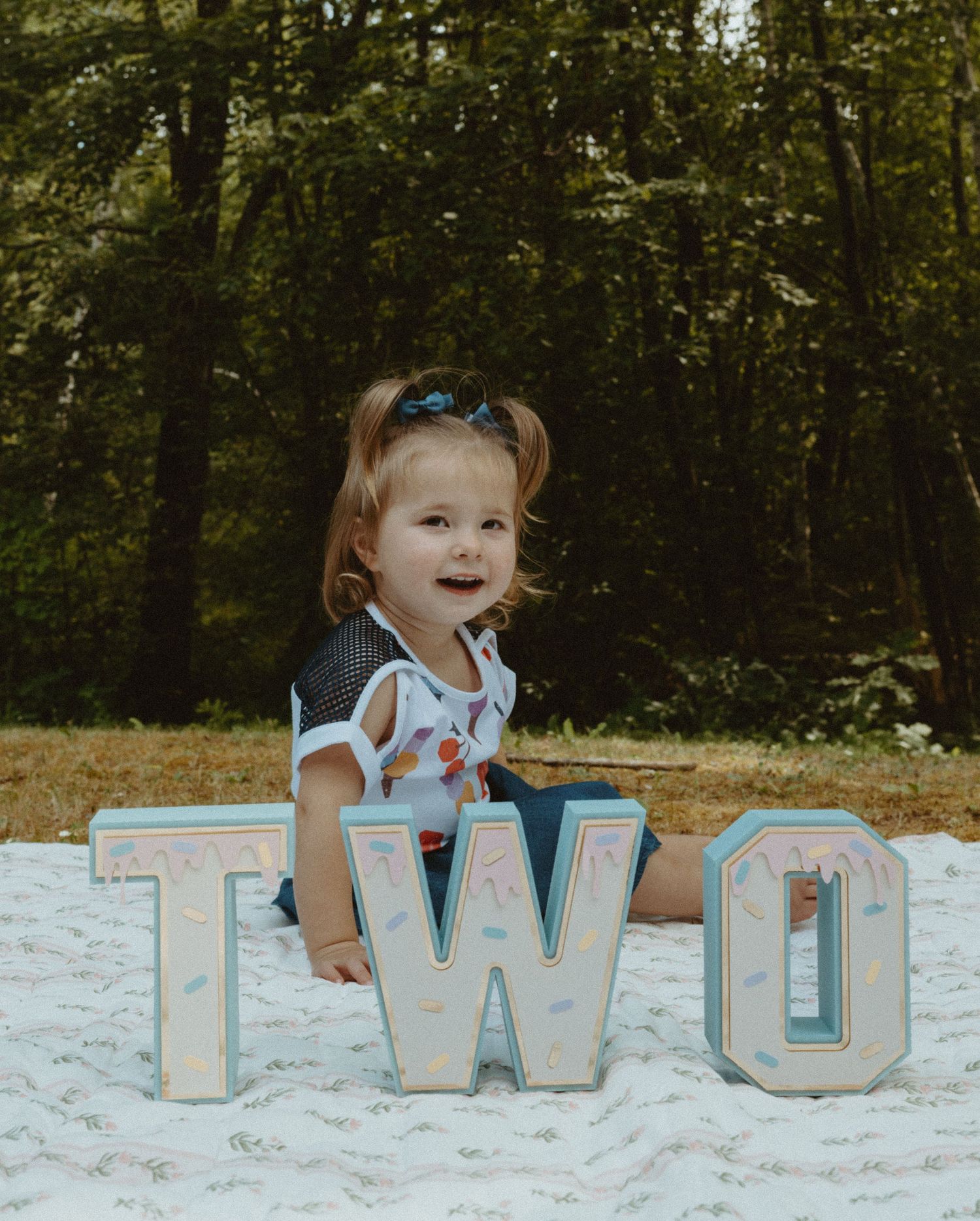 Smiling toddler girl posing with TWO letter signs for her 2nd birthday photo shoot outdoors.