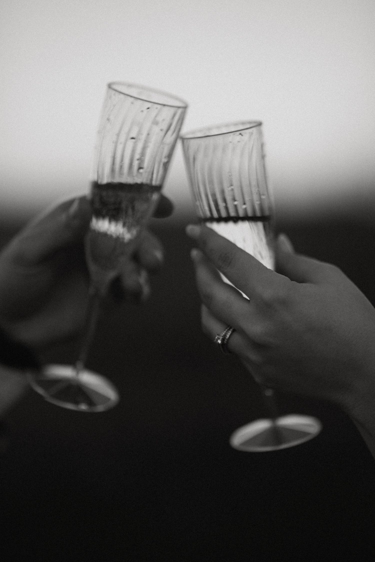 Black and white close-up of couple toasting with elegant champagne flutes, engagement ring visible.