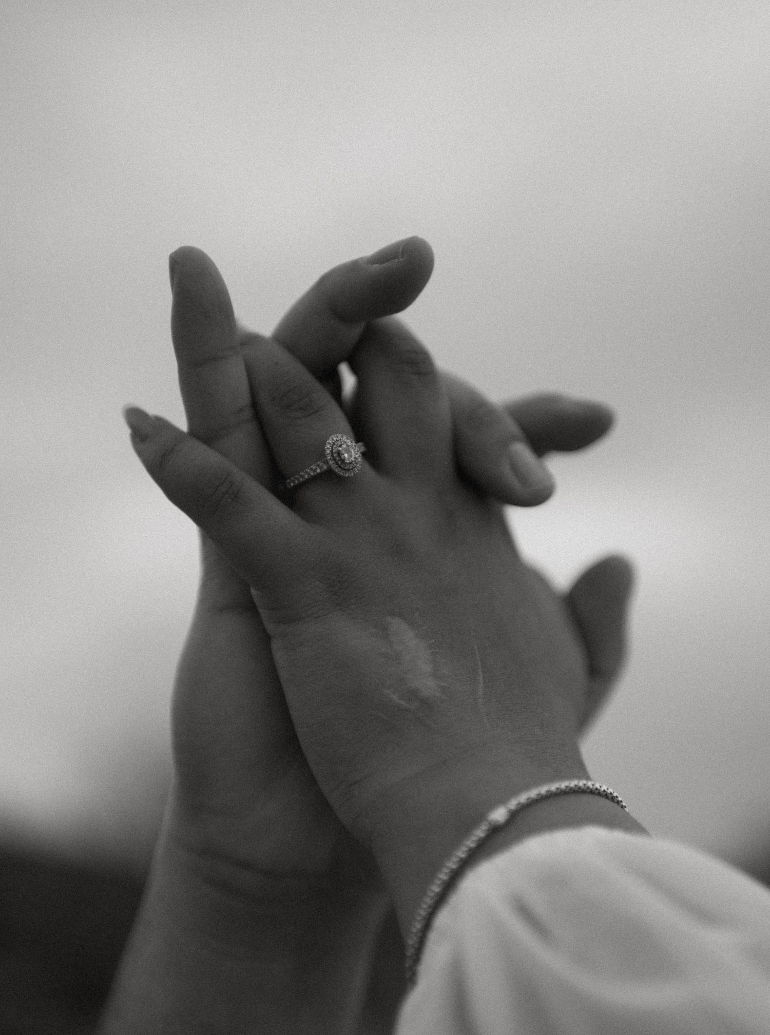 Close-up black and white photo of two hands intertwined, featuring a diamond halo engagement ring and bracelet.
