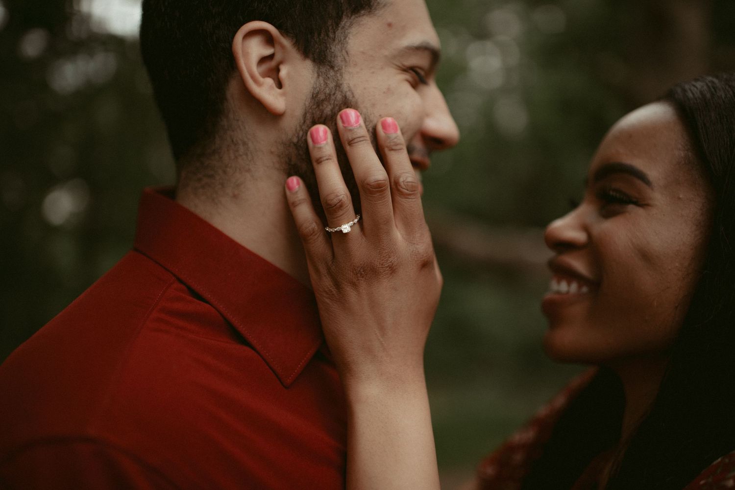 Close-up of smiling woman touching mans face with engagement ring visible on her pink manicured hand.