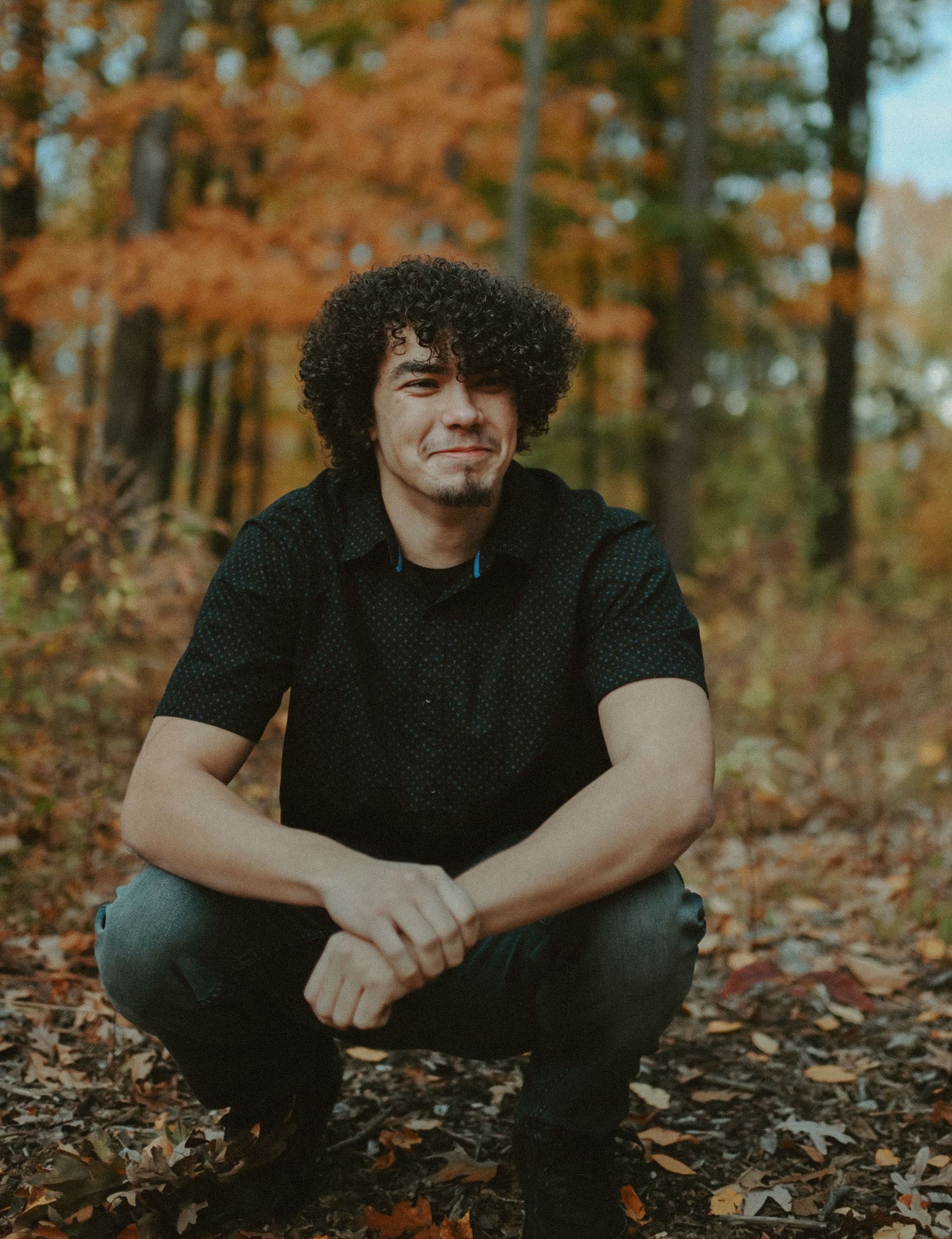 Young man with curly hair crouching in colorful autumn leaf-covered forest path.