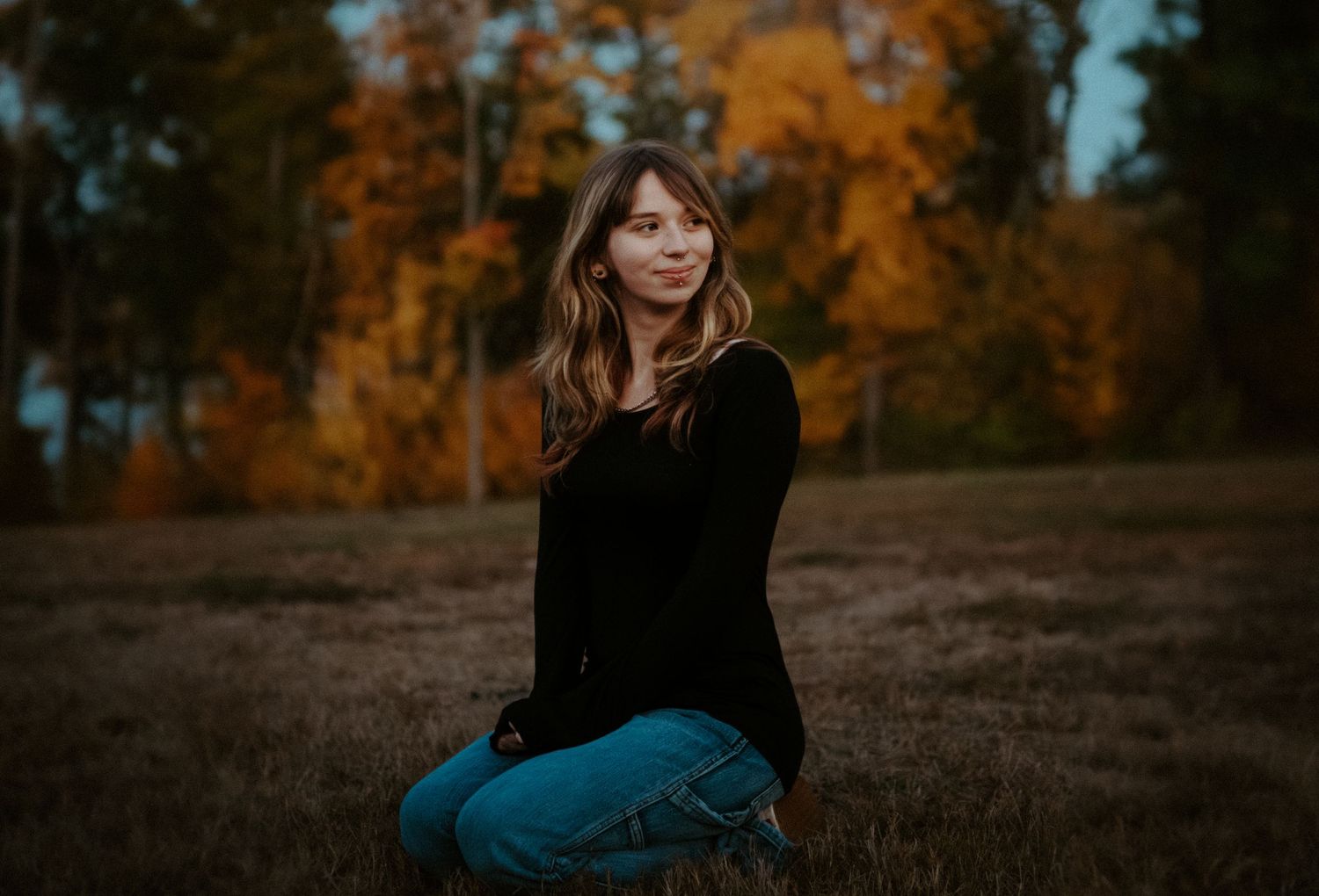 Young woman with long wavy hair sitting in an open field wearing jeans and black top with vibrant fall trees behind.