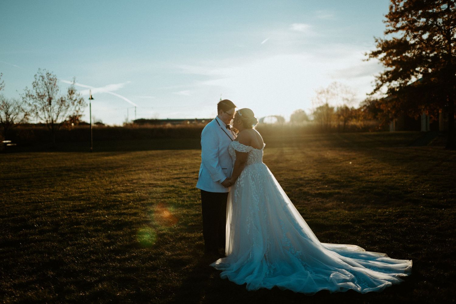 Brides embrace at golden sunset in open field, wedding dress train flowing gracefully.