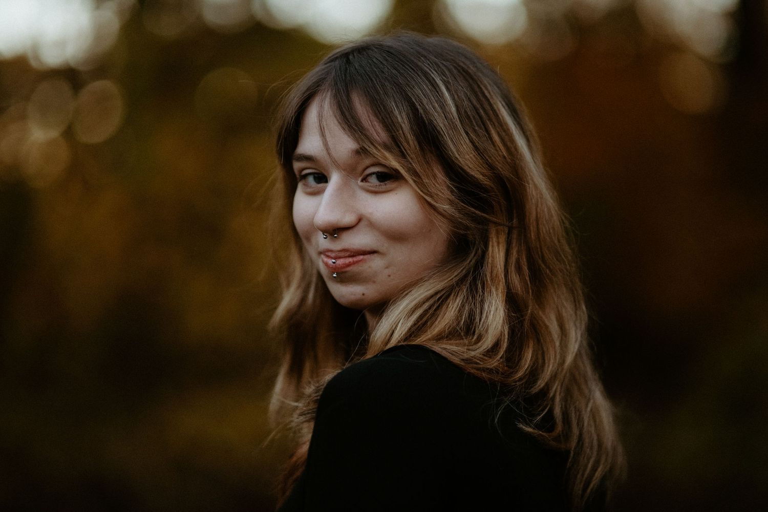 Young woman with wavy hair turning to smile over her shoulder outdoors with warm autumn bokeh background.