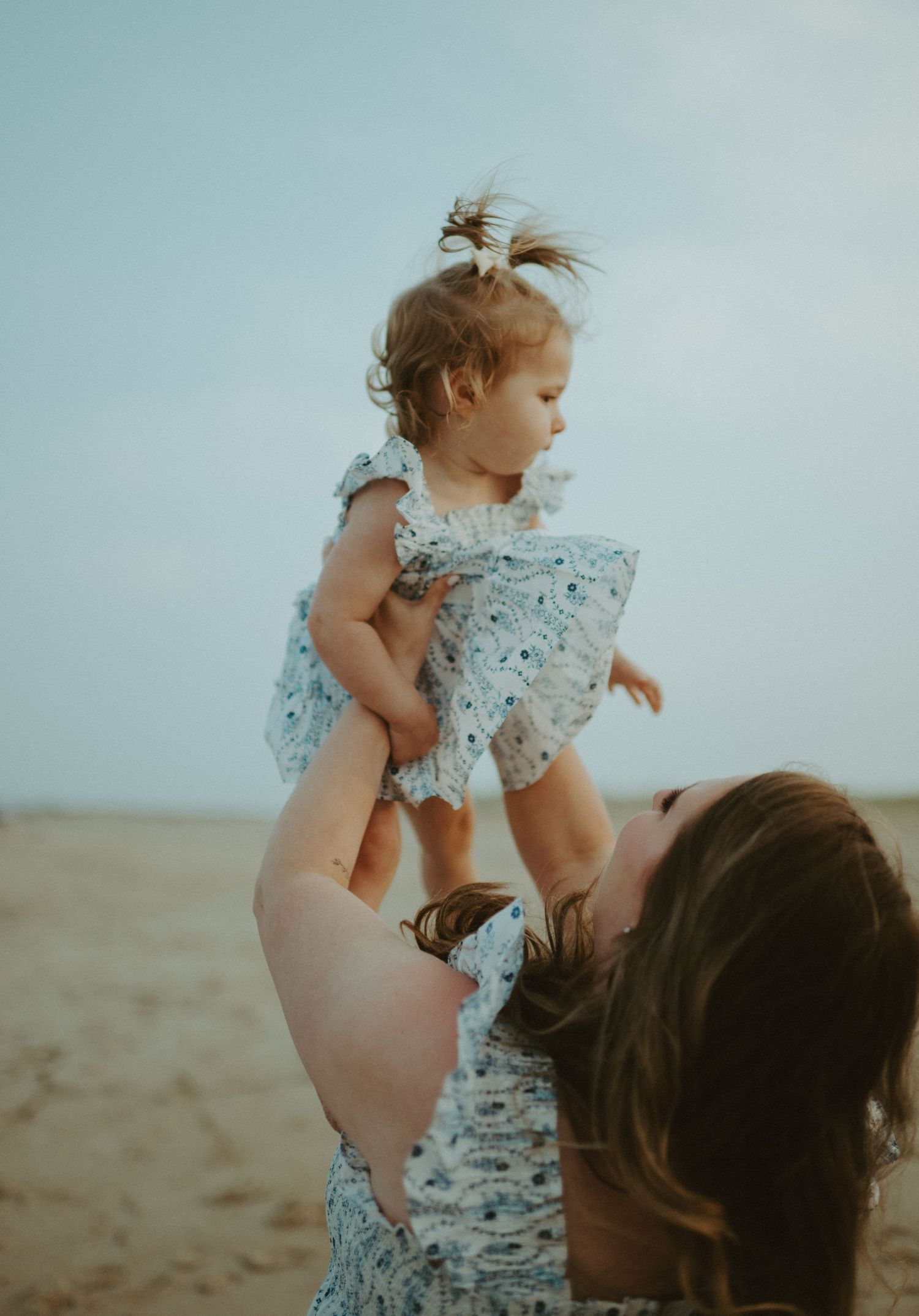 Mother lifting laughing toddler girl in floral dress high in the air on a sandy beach.