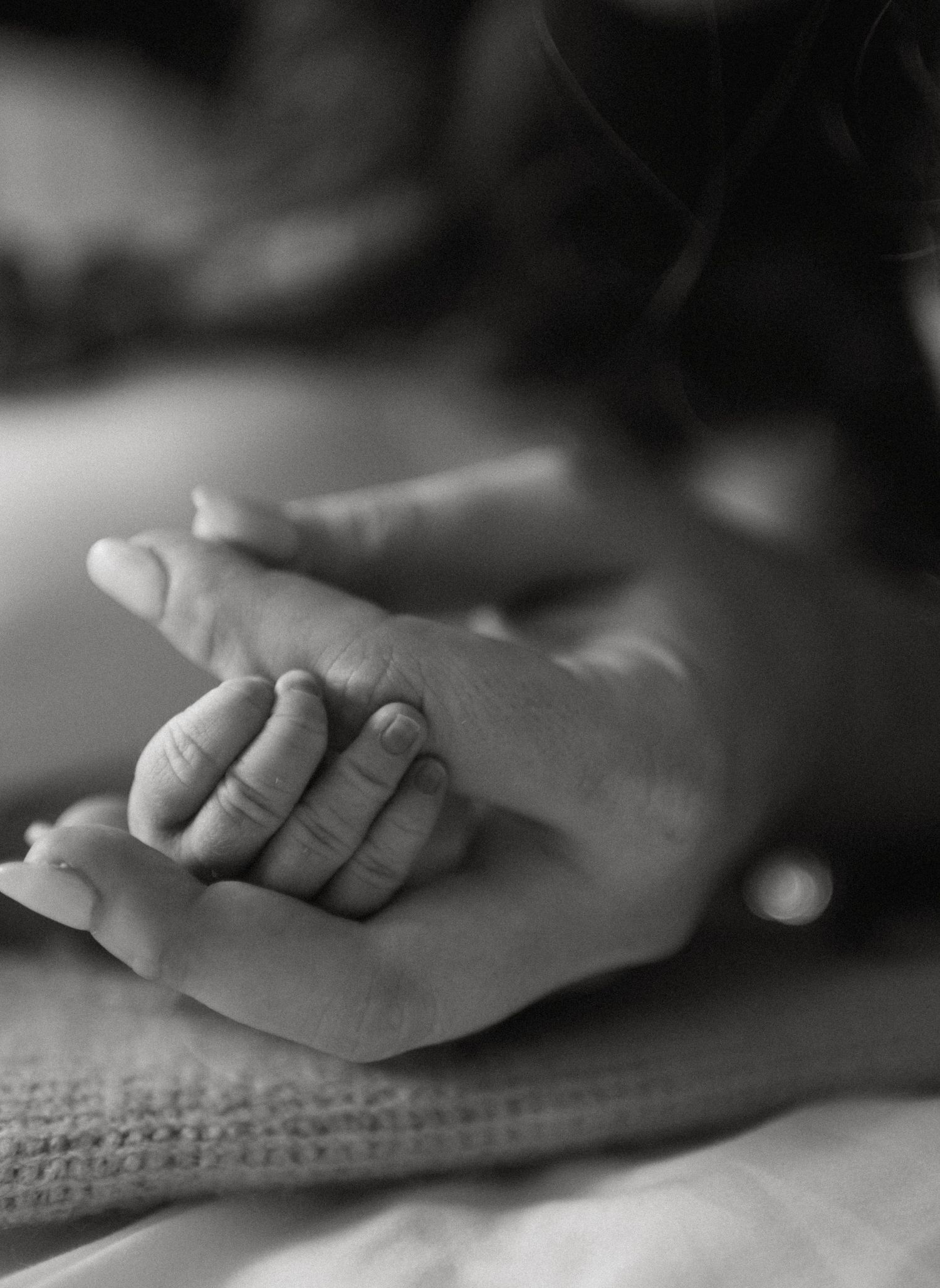 Black and white close-up of newborn baby fingers grasping mothers hand on a textured blanket.