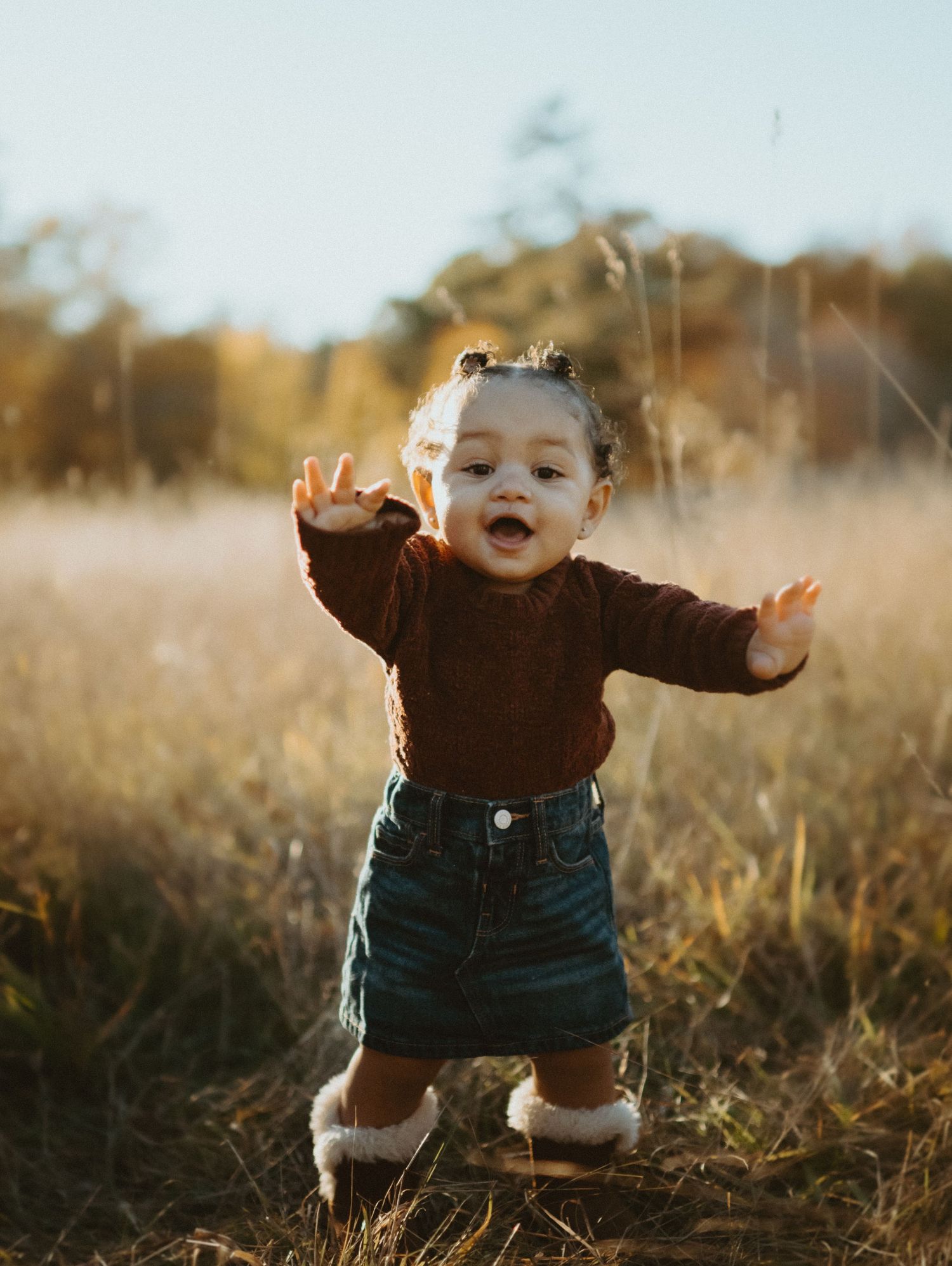 Cute baby girl in red sweater and denim skirt standing in golden autumn field with arms raised.