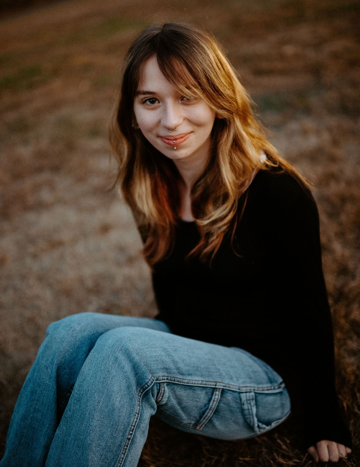 Young woman with wavy highlighted hair sitting outdoors in jeans and black top smiling softly at golden hour.