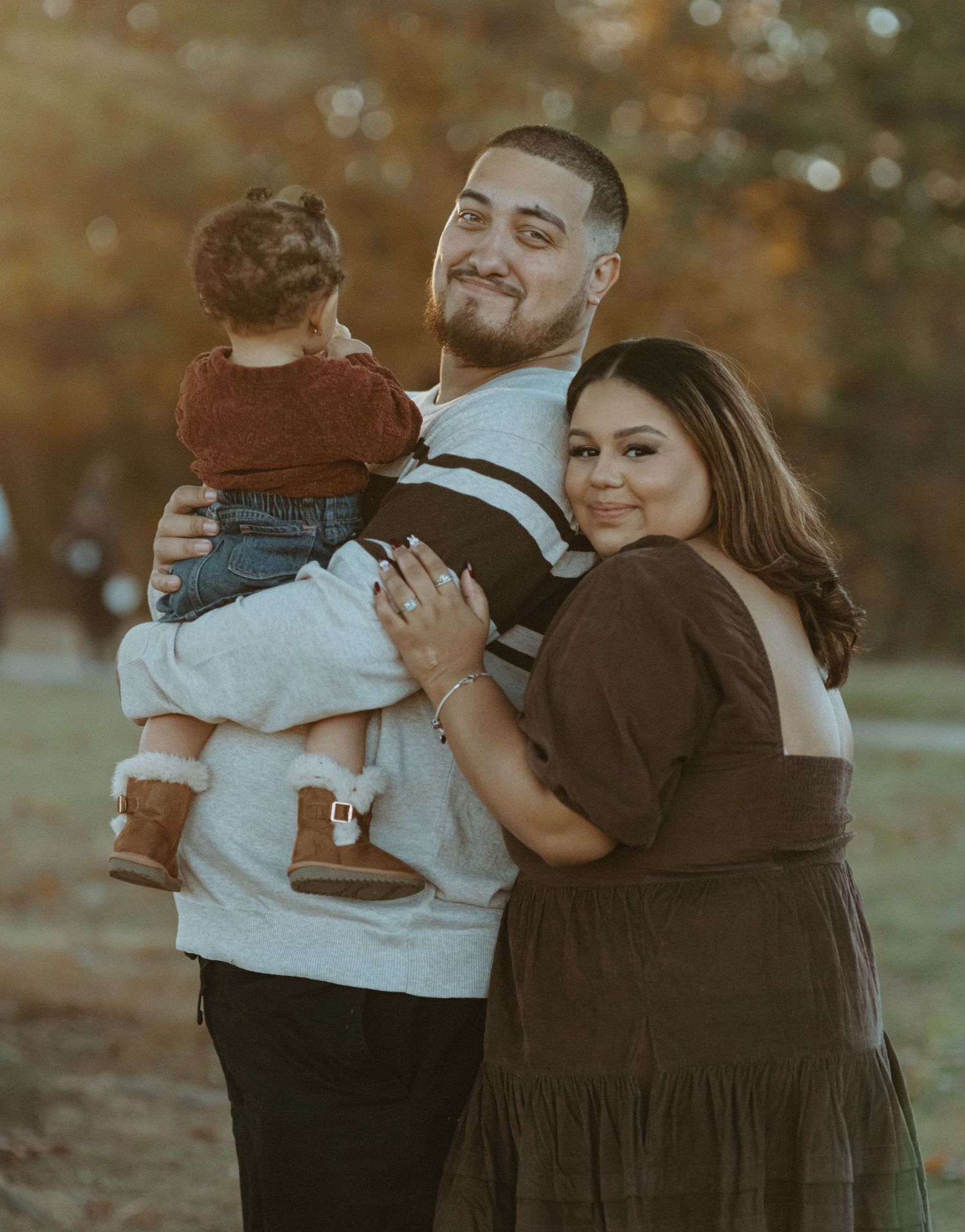 Family posing outdoors in warm autumn light, father holding toddler while mother embraces them both.